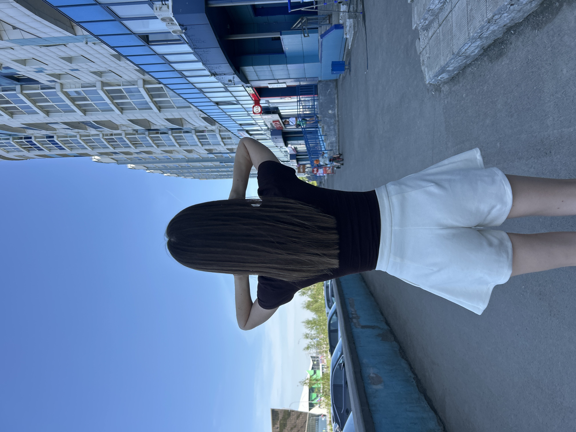 A full-length, eye-level shot captures a person from behind, standing on a paved surface next to a road, looking towards a large, modern building. The image is oriented sideways, but when rotated correctly, depicts a clear, sunny day. The subject has long, straight dark hair, wears a black short-sleeved top and white shorts, and has their hands behind their head.
In the background, a multi-story building with a facade of numerous rectangular windows and blue panels dominates the left side of the frame. Its architecture suggests a commercial or large residential complex. Below the building, near its entrance, faint figures of other people are visible.
To the right of the person, a blue curb or barrier runs parallel to the paved area, behind which several parked cars are partially visible. Beyond the cars and barrier, a line of green trees and foliage is seen under a wide expanse of clear blue sky. The bright sunlight casts distinct shadows, indicating midday.
Visible text on the building includes a red and white sign, with a clearly discernible "C&A" logo, suggesting the building houses a retail store. Other smaller, indistinct text elements are present near the building's entrance. The overall scene is an urban outdoor setting, possibly a street or parking area adjacent to a shopping center or office complex.