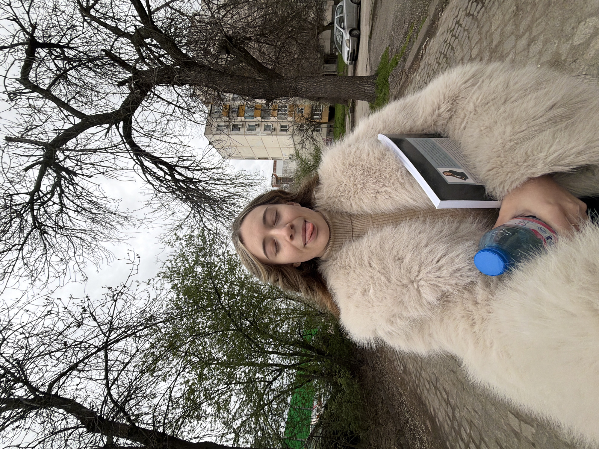 A medium shot shows a woman outdoors, possibly in Plovdiv, Bulgaria, due to the typical Soviet-era apartment block visible in the background. She is wearing a large, cream-colored fur coat, suggesting a cooler climate. She holds a book with a dark cover and a water bottle with a blue cap. The woman has light skin and blonde hair; her eyes are closed, and she sticks her tongue out slightly.
The background features bare trees and some with new green leaves, indicating early spring. There is a sidewalk or paved area where she is standing, and a car is parked further down the street. The sky appears overcast. The angle suggests the picture was taken at ground level and rotated.