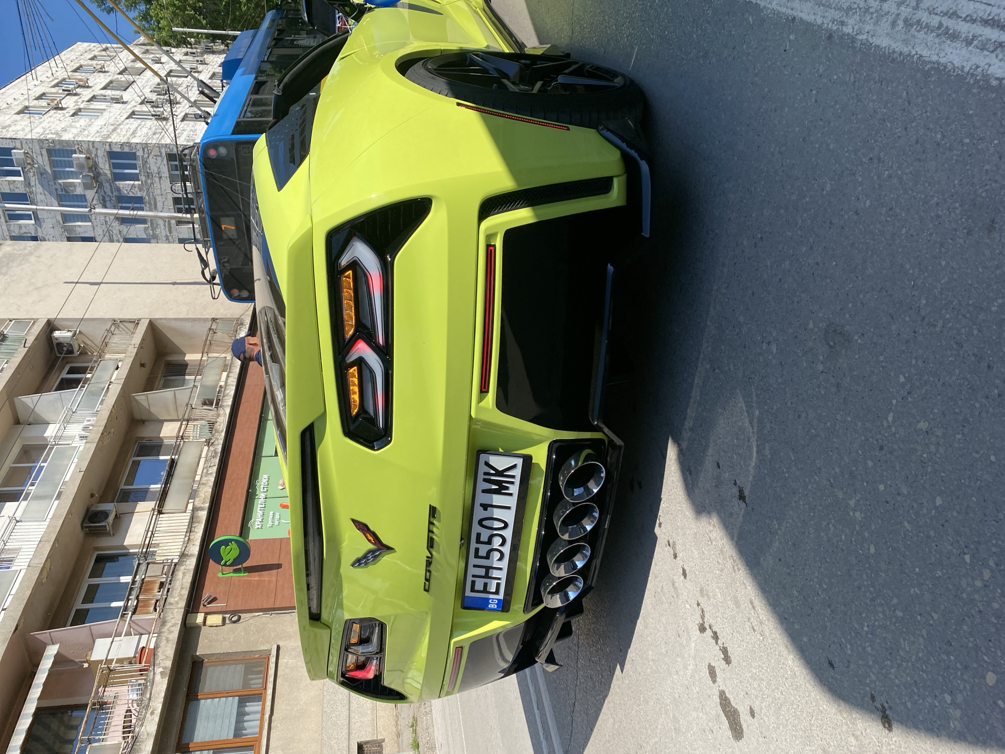 A lime-green Corvette is parked on the side of a street in Pleven, Bulgaria, casting a shadow on the asphalt. The rear of the vehicle is prominent, showcasing the quad exhaust pipes, Corvette emblem, and a Bulgarian license plate reading "EH5501 MK." Behind the car, there are buildings with architectural details, including air conditioning units and balconies. A blue bus or truck is partially visible above the Corvette. The buildings suggest a typical urban street scene in Bulgaria.