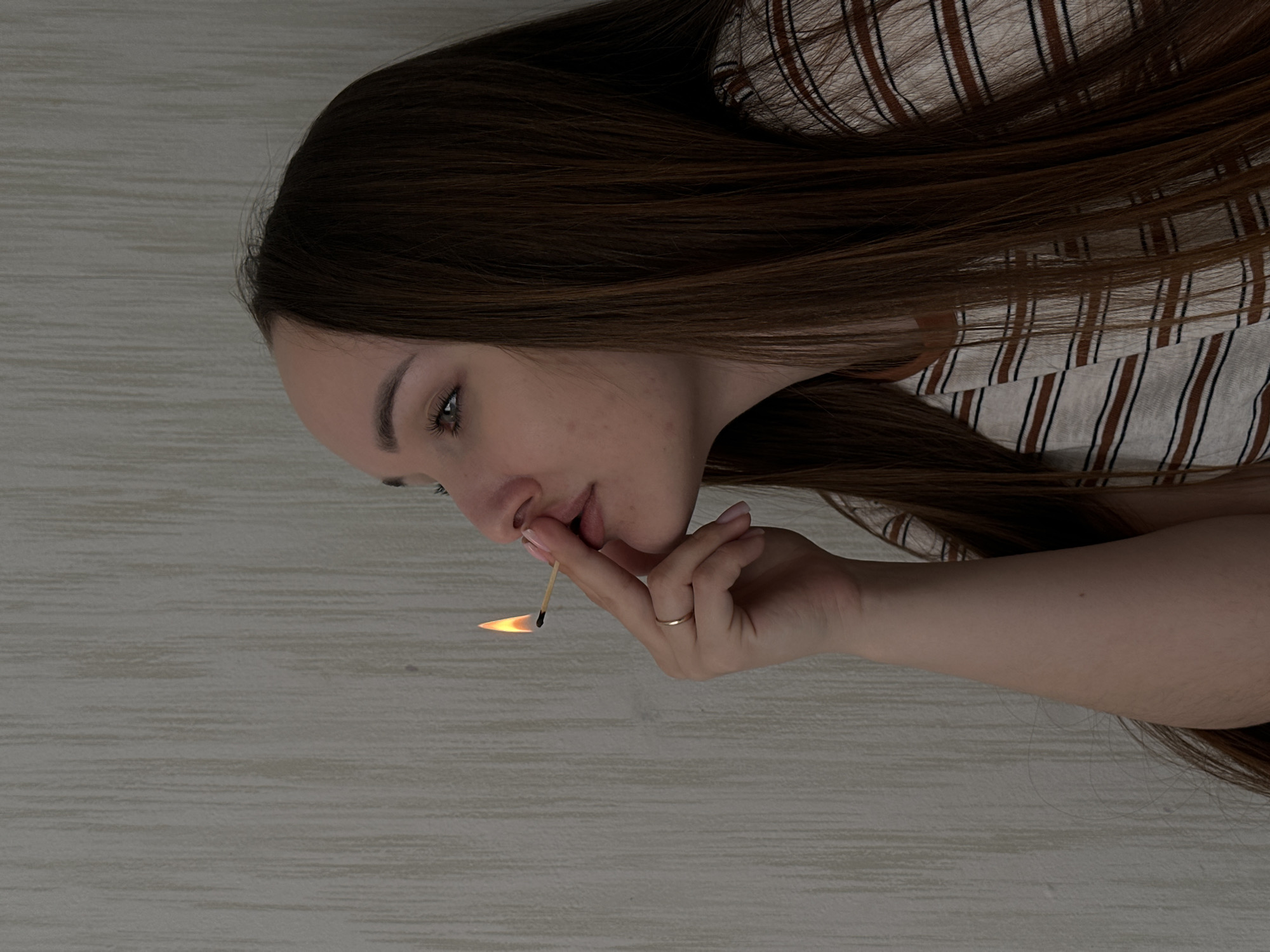 A woman with long brown hair is holding a lit matchstick to her lips. She has fair skin, a gold ring on her finger, and wears a striped shirt. The background is a light-colored surface that appears to be a wood floor or countertop. The location cannot be verified as Chaikovsky, Russia, based solely on this image.