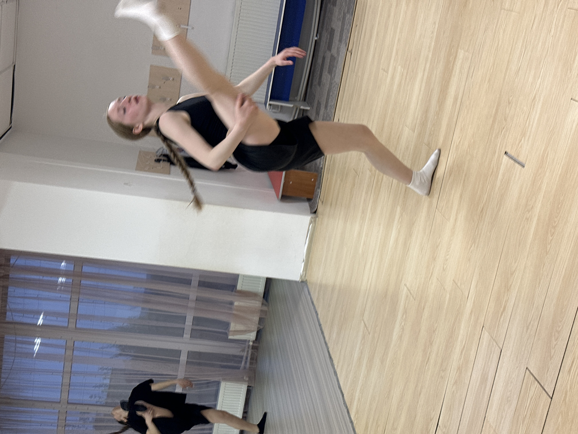 The image shows two female dancers in a dance studio. The primary dancer is in mid-air, executing a high leg extension while holding her extended leg with her hand. She is wearing a black leotard and black shorts, with white socks. Her blonde hair is braided. In the background, a second dancer is on the floor performing a different pose. The studio has a wooden floor and large windows with sheer curtains. The location is consistent with a dance studio environment, likely in Votkinsk, Russia, given the request. The scene captures a dynamic moment in a dance session.