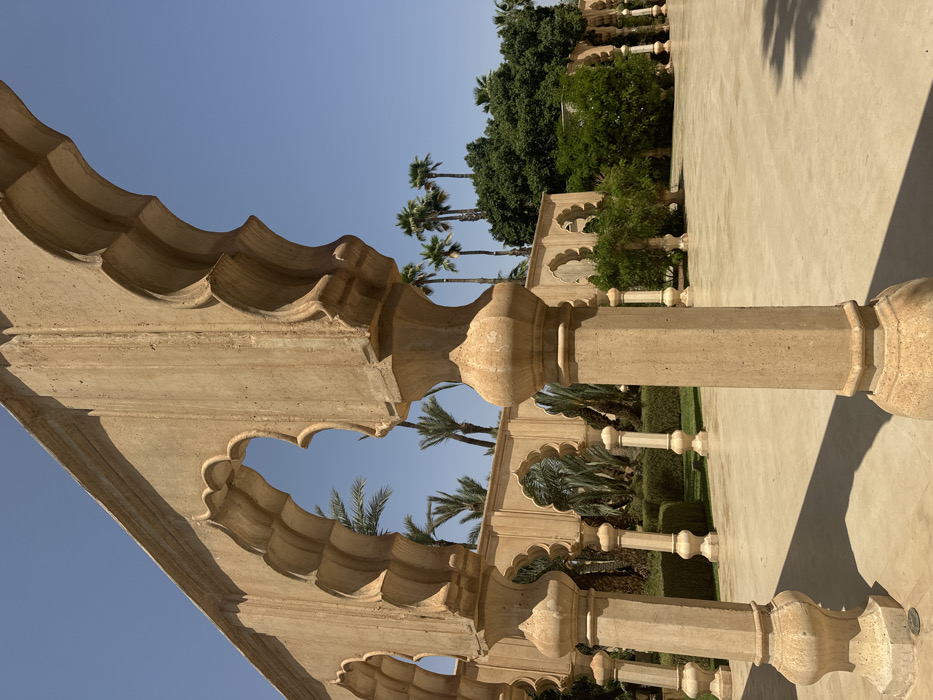 The image shows an arched walkway lined with stone pillars in a courtyard in the city of Ouled Jelal, Morocco. The pillars are all made of a light tan stone and are adorned with intricate carvings. The walkway leads to a paved courtyard. There are palm trees and other greenery lining the courtyard and the walkway. The sky is blue and cloudless. The image was taken from the perspective of someone standing under one of the arches, looking toward the courtyard. The image was taken at a location with coordinates latitude: 31.663, longitude: -7.872.