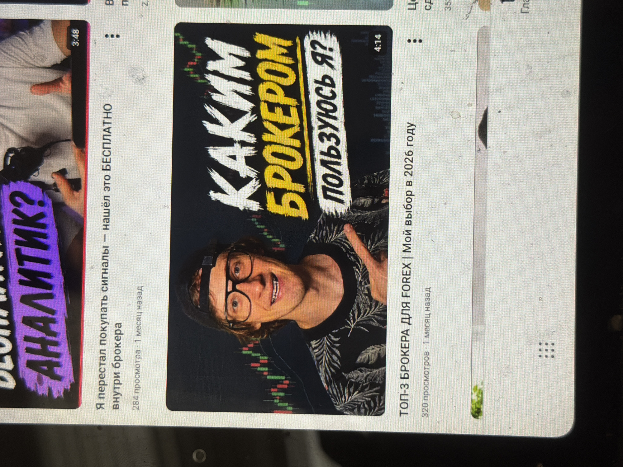 The image shows a screenshot of a YouTube interface displaying several video thumbnails. The primary focus is on a video with the title "КАКИМ БРОКЕРОМ ПОЛЬЗУЮСЬ Я?". The thumbnail features a man wearing glasses and a black shirt with a floral pattern, pointing to the right. The video is 4 minutes and 14 seconds long and has 320 views, posted 1 month ago. Above this, another video is partially visible with the text "АНАЛИТИК?" and a title that includes "Я перестал покупать сигналы". To the right, another video thumbnail shows the text "В" and "п". Further down, there's a partial view of another video with the title "Ц" and "сд". The overall environment appears to be indoors, with the screen of a device displaying the YouTube content. There are no discernible elements that would indicate a specific city or location.