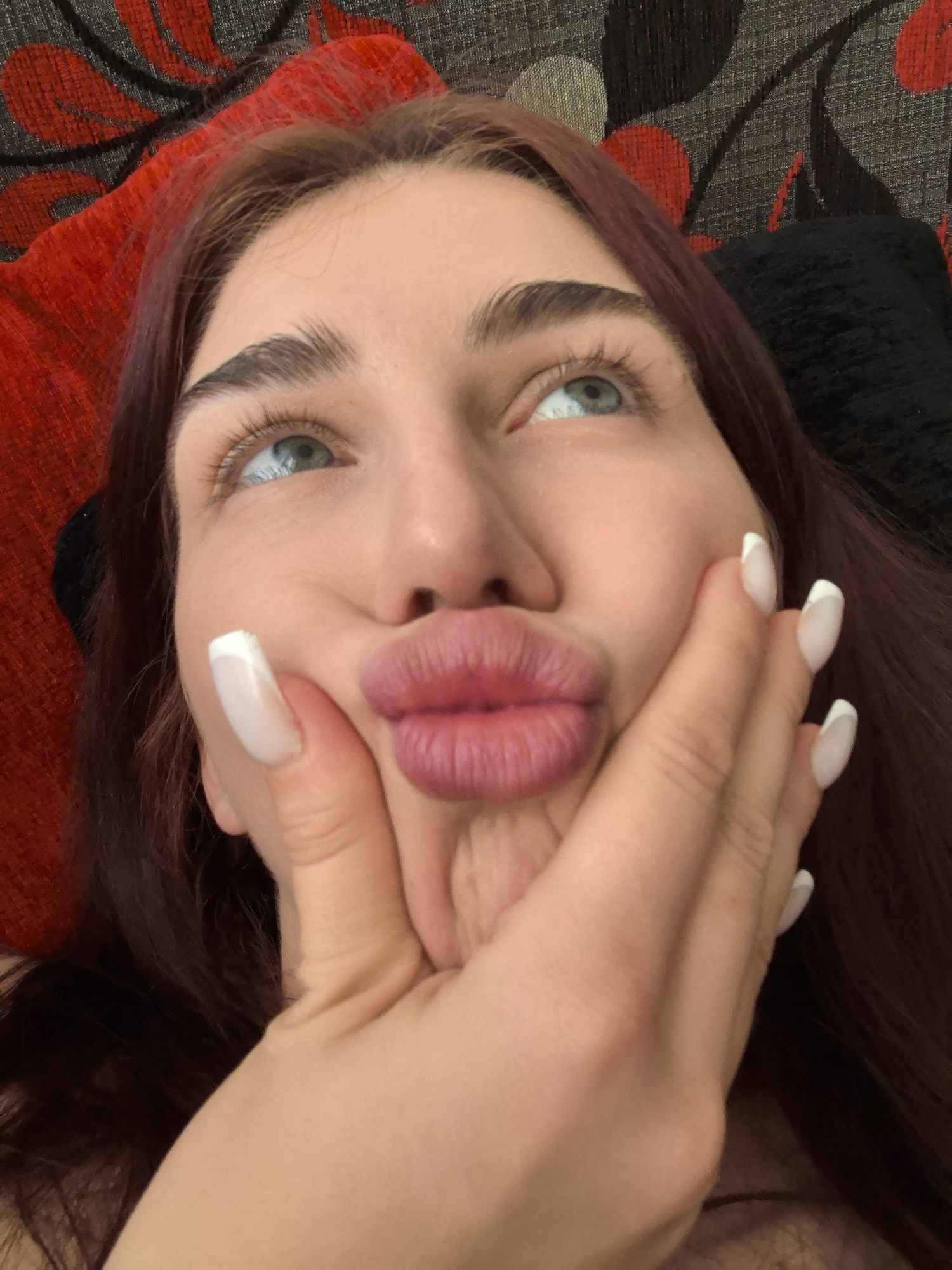 A close-up, low-angle, indoor photograph shows a young woman with auburn hair and light blue eyes looking upwards. Her cheeks are pressed inwards, and her lips are pursed in a duckface pose. Her right hand is raised, with her thumb and index finger pinching her cheek, and her fingernails are painted with white French tips. She is resting against a textured surface that appears to be a couch or upholstered chair, with a red and black patterned fabric visible behind her. The lighting is soft and appears to be artificial, casting subtle shadows on her face. There is no information in the image that suggests a location in Stara Zagora, Bulgaria.