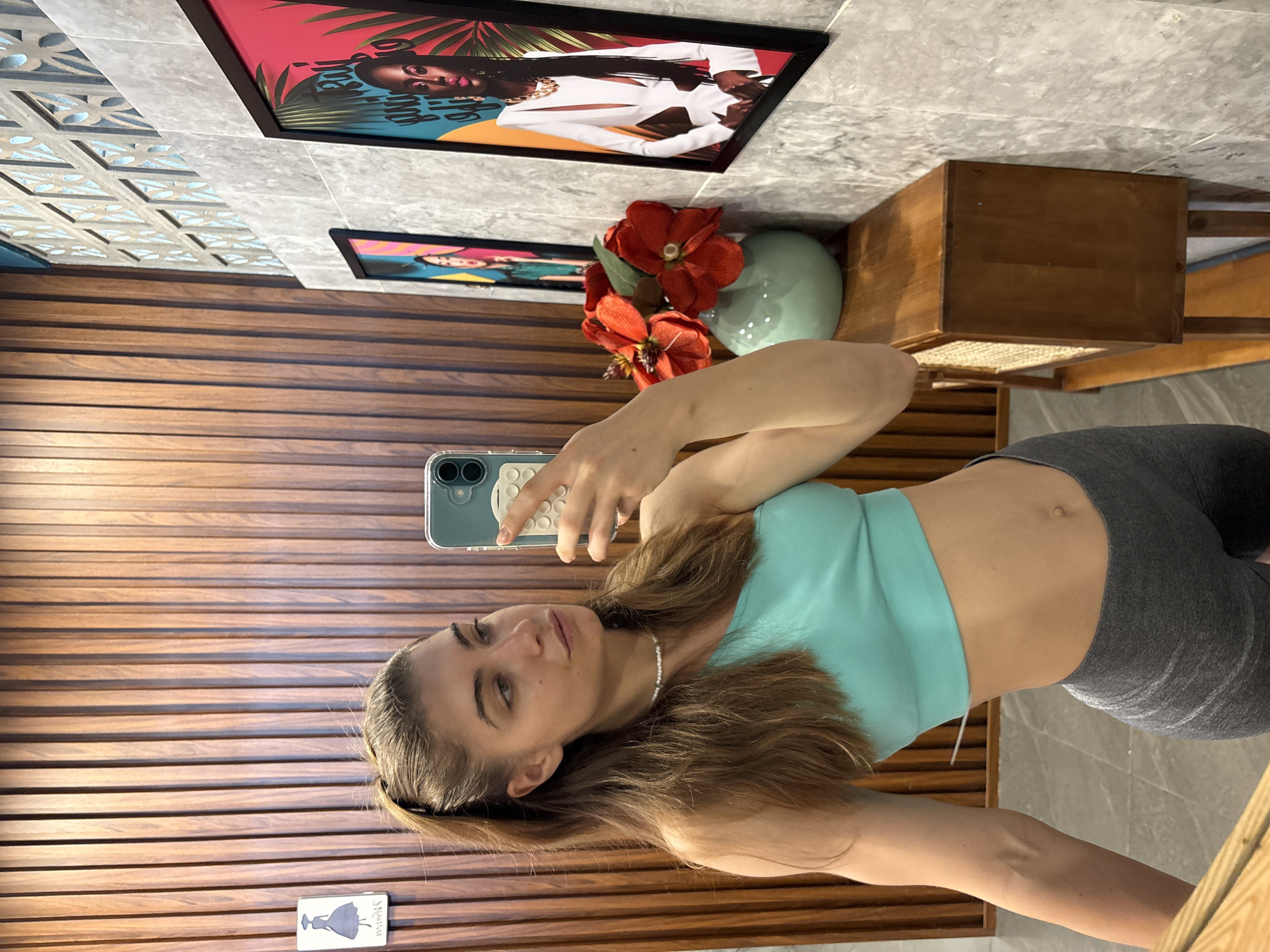 A young woman is taking a selfie in what appears to be a bathroom or spa-like setting. She is wearing a light blue crop top and gray leggings. Her hair is brown and she has a headband on. She is holding a smartphone in her left hand, taking the photo. The room has a wooden slatted wall behind her. Above the wooden wall, there is a section of patterned tile. There are also framed pictures on the wall, one of which depicts a woman in a white blazer. There is also a small wooden cabinet or side table with a vase of red flowers on top. There is a bathroom sign is visible near the bottom of the frame. Without additional clues, it is not possible to confirm the location in Đà Nẵng, Vietnam.