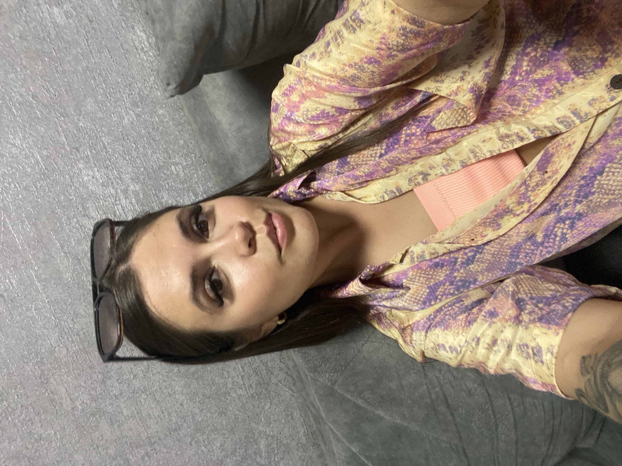 A woman with brown hair and makeup, wearing sunglasses on her head, a patterned shirt, and a pink top, is photographed from the chest up, looking at the camera. She is positioned in front of a gray textured wall and a gray couch. The image was taken in Makhachkala, Russia.