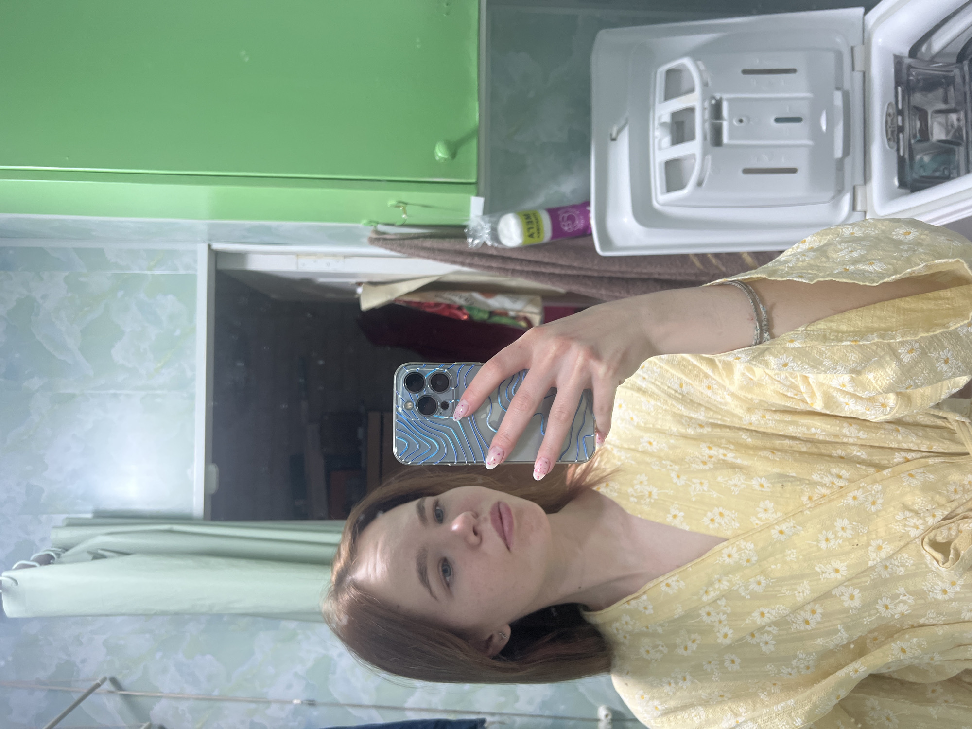 A fair-skinned woman with light brown hair is taking a selfie in a bathroom. She is wearing a yellow floral print top. She is holding a smartphone with a blue topographic case. The bathroom has green cabinets and pale marble-patterned walls. There is a small white cabinet open above the woman’s right shoulder. This location is in the city of Chaikovsky, Russia.