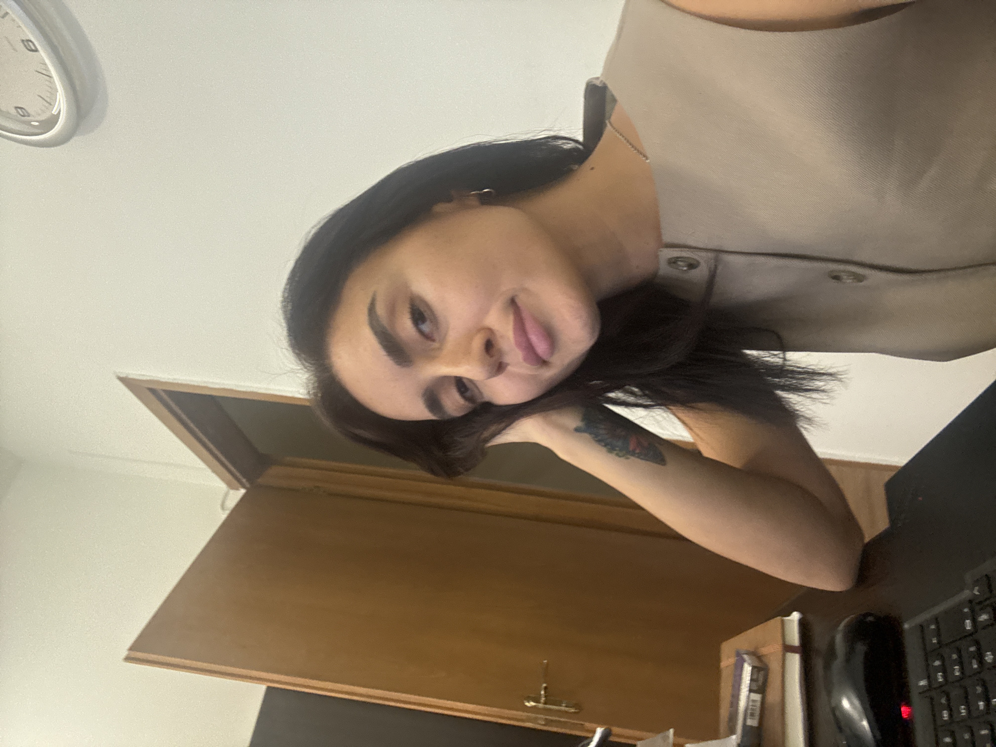 A young woman with dark hair and light skin, wearing a light-colored vest, rests her head on her hand and looks towards the camera. She is seated at a desk with a keyboard and mouse in front of her, and a notebook and pen to her left. Behind her, a wooden door is slightly ajar, revealing a glimpse of a dark interior. A clock hangs on the wall to her right. The scene appears to be an indoor office or study.