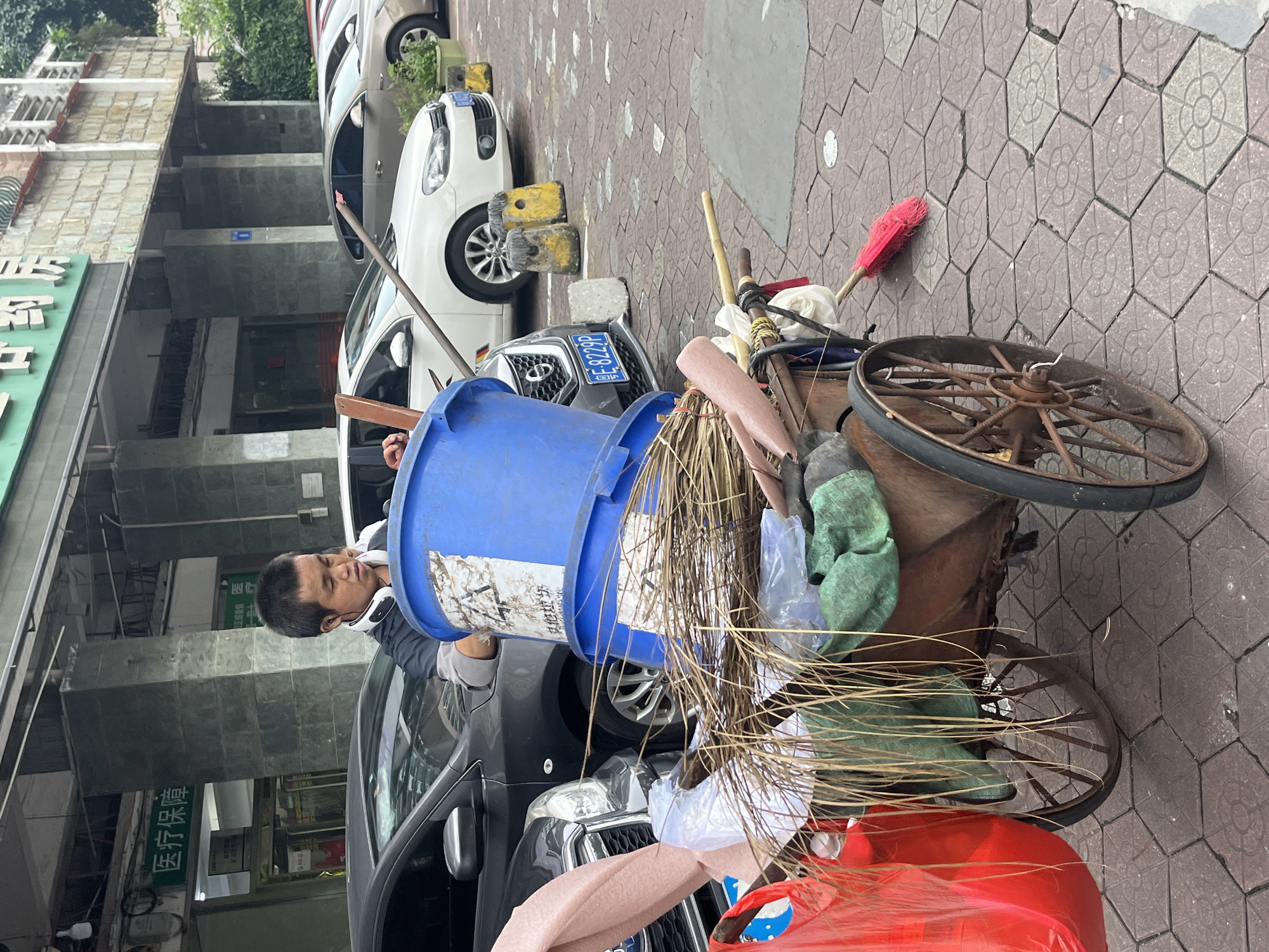 A male sanitation worker, appearing to be in his late 20s or early 30s, stands beside a two-wheeled cart laden with cleaning supplies. He wears a dark-colored shirt with light gray sleeves and headphones around his neck. The cart is filled with two large blue buckets, one stacked atop the other, and an assortment of brooms, dried plant material, and plastic bags. A red plastic bag is visible on the left side of the cart, and a red-bristled broom lies on the tiled ground to the right. Behind the cart, several cars are parked, including a dark gray sedan with a license plate reading "E 8229P" and a white hatchback. The scene takes place on a paved walkway in front of a building with a green sign that reads "医疗保障" (Medical Security). The building's facade is constructed with stone tiles, and vertical pillars support an overhang. The area appears to be an urban setting in Chancheng District, China, likely in the vicinity of a medical facility.
