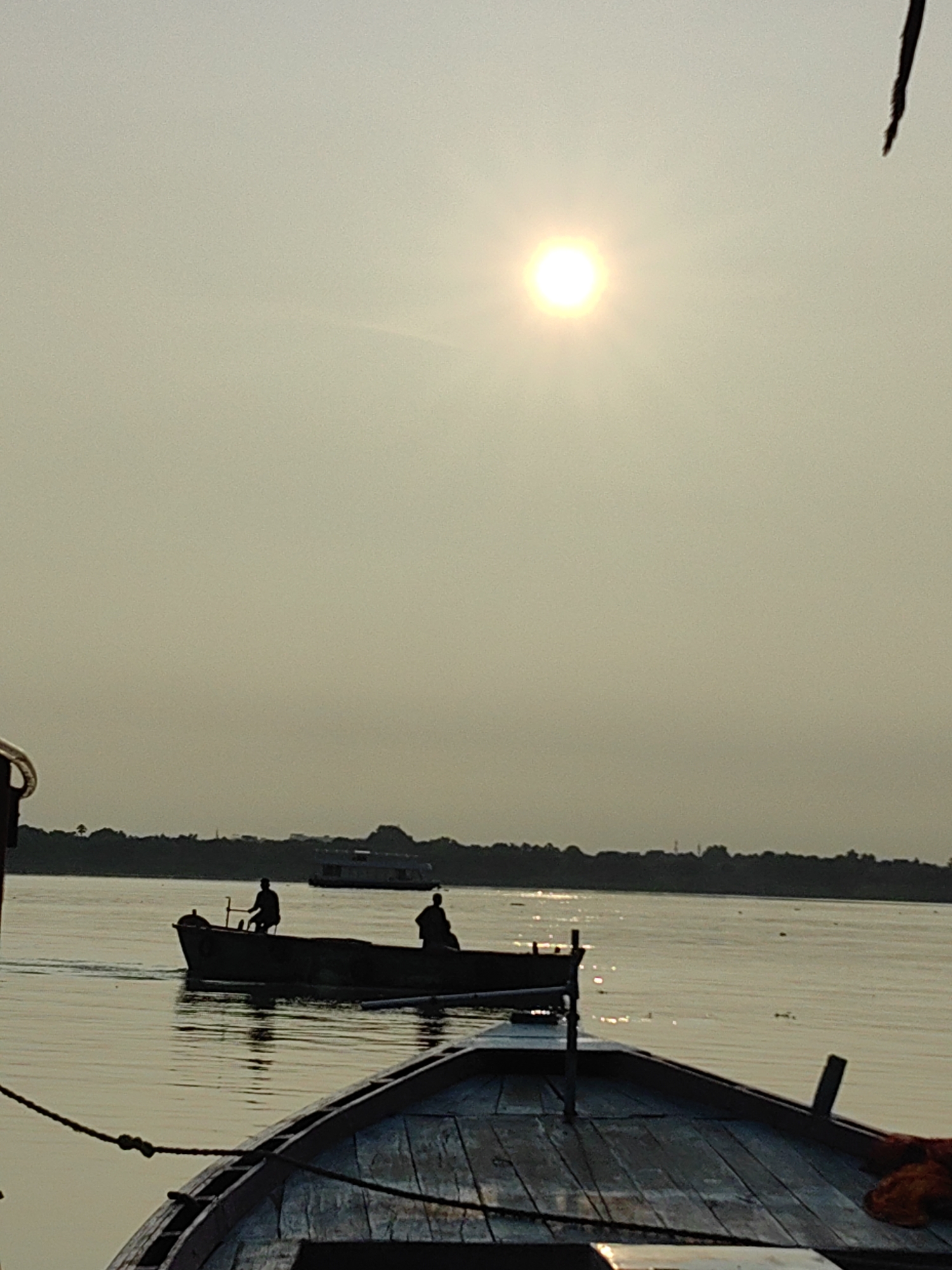 This is a vertical, medium-shot photograph taken from a boat on the Ganges River in Varanasi, India, during sunset. The sun, a bright, circular orb, is positioned in the upper right quadrant of the sky, casting a warm, hazy glow. A silhouetted boat with two figures is visible in the middle ground, moving from left to right across the river. The boat in the foreground, from which the photo is taken, is also silhouetted, with its wooden deck and some ropes visible. A dark line of trees and buildings forms the shoreline in the background. The overall atmosphere is serene and tranquil, with the water reflecting the soft light of the setting sun.
