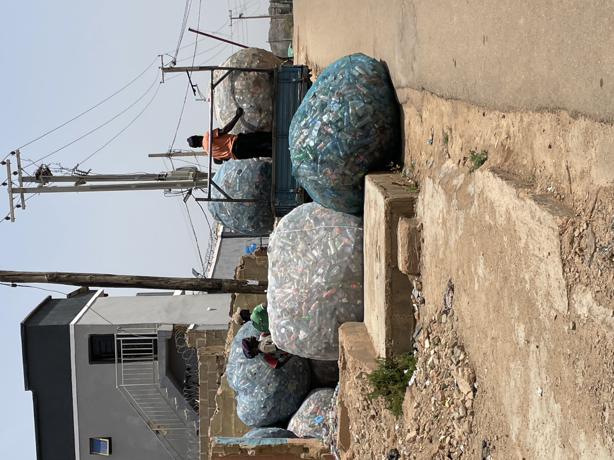 The image depicts a scene in the city of Jos, Nigeria, showcasing a collection of large bags filled with plastic bottles. These bags are situated on the side of a road, with some positioned on the truck. An individual is standing on the truck, seemingly organizing or inspecting the bags. Other people are also visible among the bags, possibly involved in collecting or sorting the bottles. The environment includes buildings, power lines, and a dirt and concrete terrain characteristic of an urban or peri-urban area in Jos. The scene reflects recycling or waste management activities taking place in the city.
