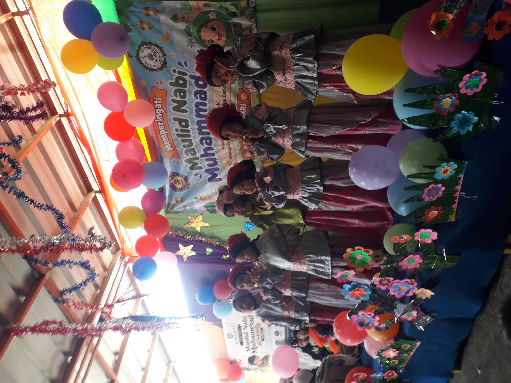 In Cankuang, Indonesia, a group of young girls, dressed in matching grey tunics with pink trim and matching headscarves, stand on a decorated stage. They are participating in an event commemorating Maulid Nabi Muhammad SAW. Some of the girls pose for the camera with peace signs, while others look more candidly towards the audience. The stage is adorned with colorful balloons in shades of pink, purple, yellow, and blue, as well as decorative tinsel garlands hanging from the ceiling. A banner behind the girls reads "Memperingati Maulid Nabi Muhammad SAW," indicating the purpose of the gathering. The backdrop of the stage is a green curtain, and at the far right, a cartoon illustration of a girl in a hijab holding a book is visible. The overall atmosphere suggests a festive and celebratory occasion for the local community.