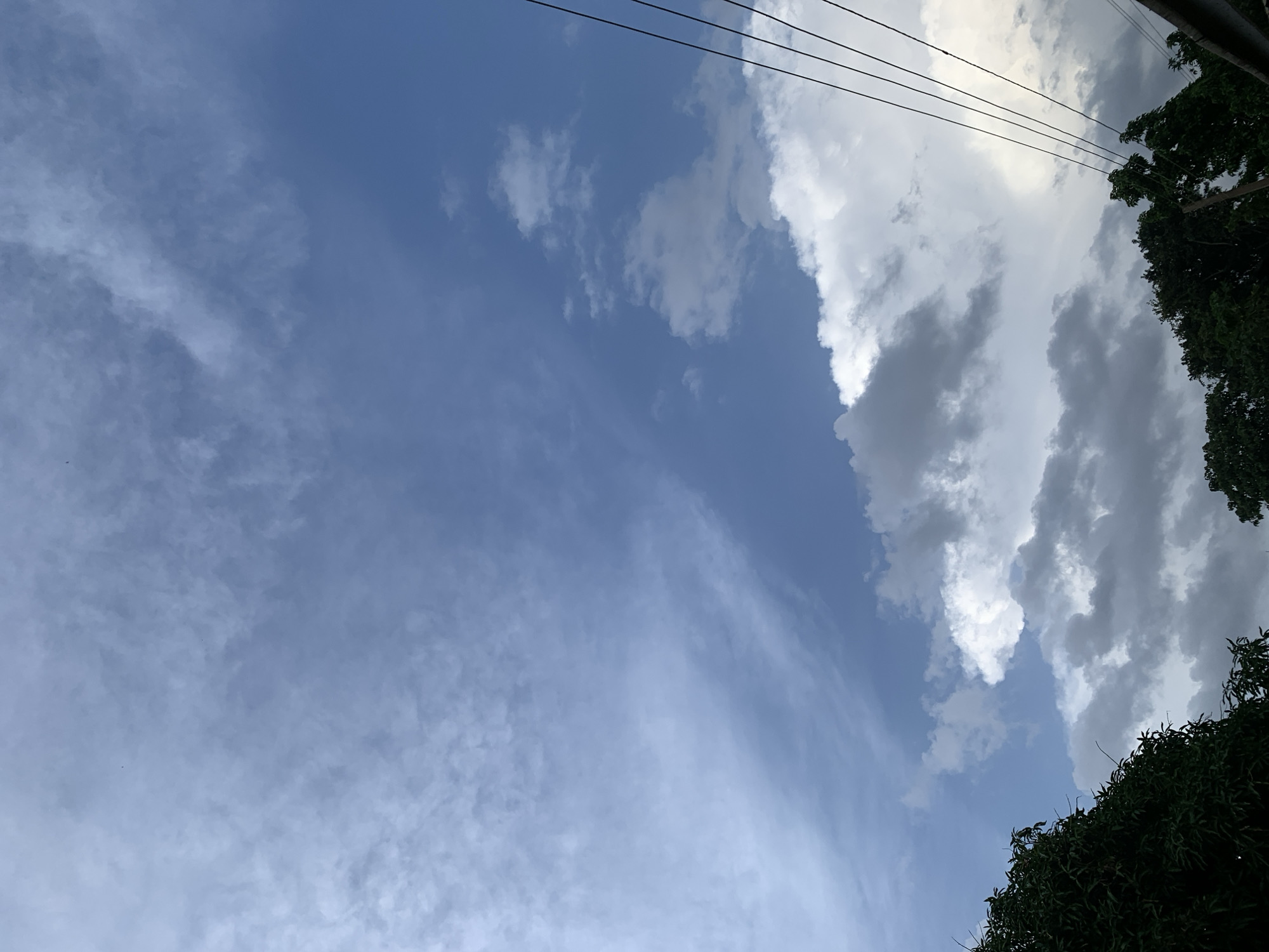 The image depicts a cloudy sky over Kuru Karama, Nigeria. The sky is a mix of light and dark clouds against a blue backdrop. In the foreground, the tops of green trees are visible on the left and right sides. Several dark electrical wires run diagonally from the upper right to the lower right corner of the frame. The scene appears to be an outdoor environment, likely during the daytime given the natural light.