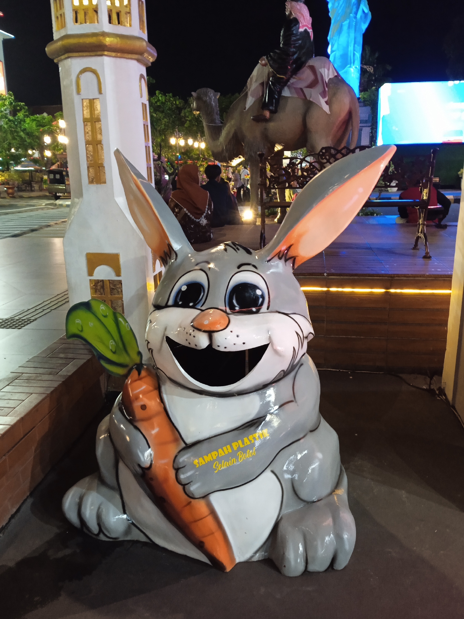 The image depicts an outdoor night scene in Madiun, Indonesia, featuring a large, cartoonish gray and white rabbit statue holding a carrot. The rabbit statue is likely a trash receptacle with "SAMPAH PLASTIK" (plastic waste) printed on it. Behind the rabbit, a white architectural structure with gold trim stands to the left, while a camel statue with a person on its back is visible in the center background. Several people can be seen walking in the background, and a brightly lit platform or stage is present. The overall atmosphere is illuminated, suggesting an urban public space in the evening.