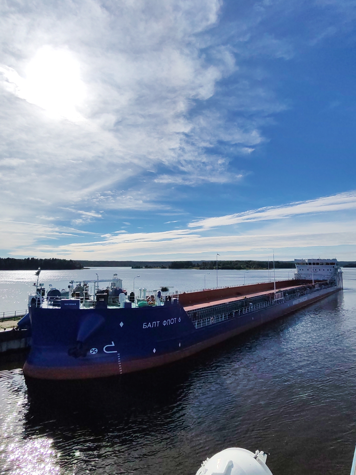 A long, dark blue barge with a reddish-brown cargo hold is docked at a pier. The barge is named "БАЛТ ФЛОТ 6" in white lettering. The bow of the barge is visible in the foreground, featuring a large anchor and a chain. The vessel is on a body of water with a clear blue sky above, dotted with wispy white clouds. A bright sun is visible in the upper left corner of the image. The background shows a tree-lined shore. The scene is likely located in Анхимово, Russia, given the context.