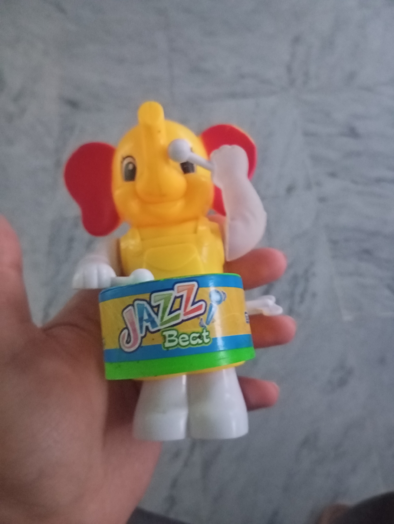 A plastic yellow elephant toy with red ears and a white stick in its trunk is held in a person's hand. The elephant is wearing a yellow shirt and blue pants, and it is standing on a cylindrical drum with the words "JAZZ Beat" written on it. The drum is yellow with blue and green stripes. The background is a gray, textured surface, possibly a floor or wall.