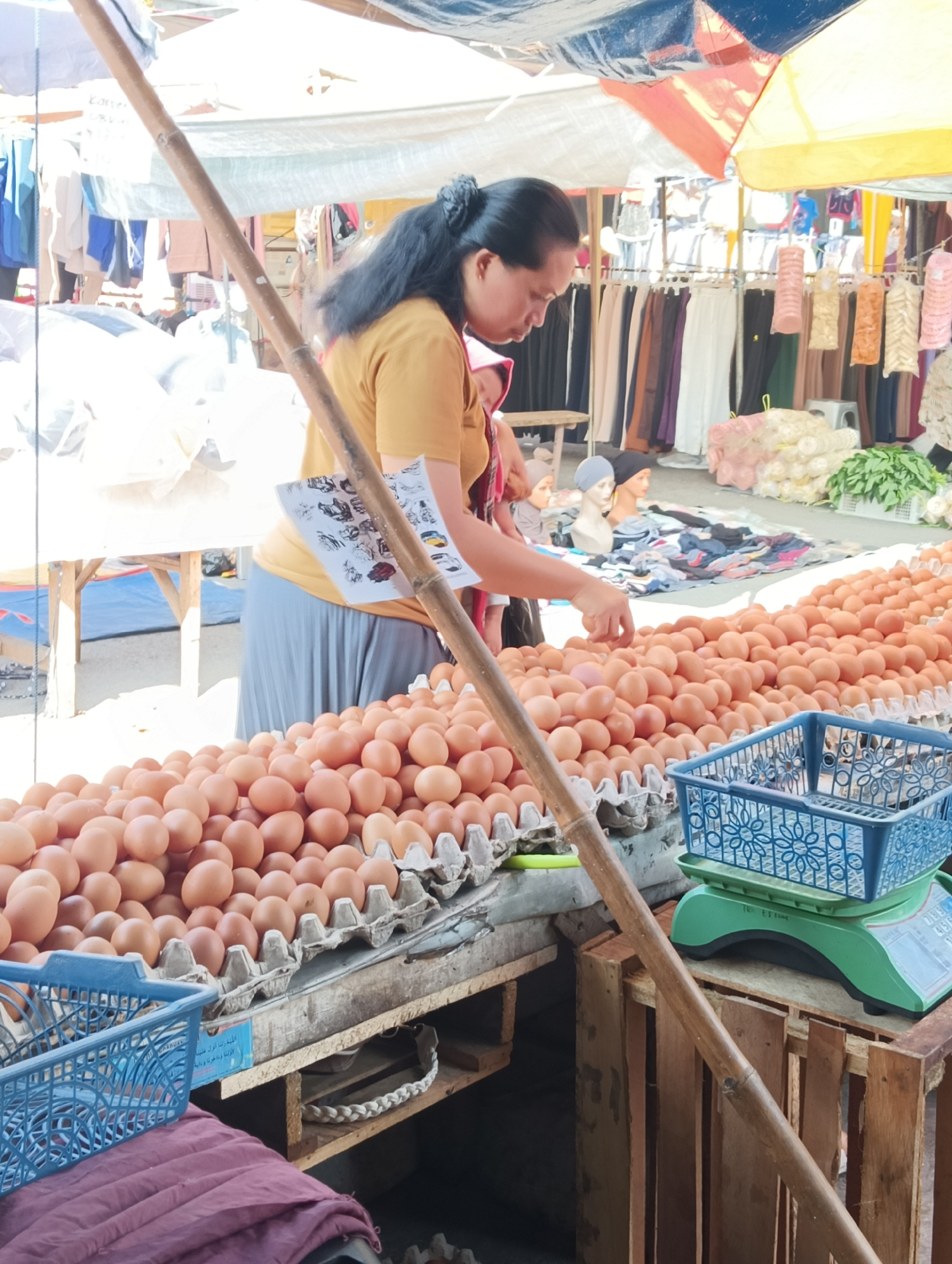 An outdoor market scene in Katapang, Indonesia, features a woman in a yellow shirt and gray skirt attending a stall filled with a large quantity of brown eggs. She is reaching into the eggs, possibly for selection or arrangement. Several rows of eggs are visible in cardboard cartons and a blue plastic basket. A bamboo pole runs diagonally across the foreground, obscuring part of the stall. In the background, a variety of goods are displayed, including clothing on hangers and bundles of fabric. A green scale with a blue basket sits to the right of the egg display. The market appears to be covered by a colorful canopy.