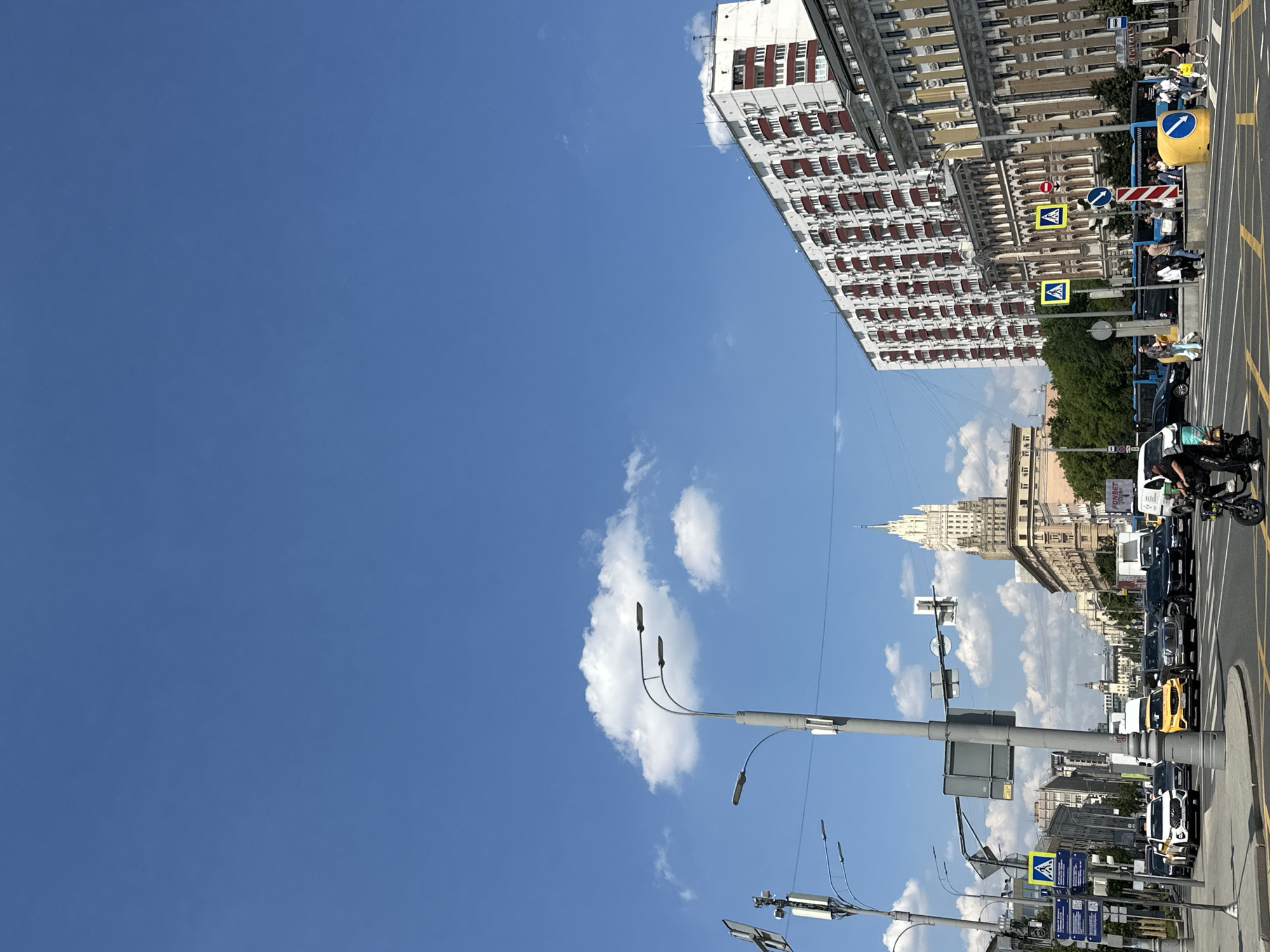 The image depicts a street scene in Moscow, Russia, likely on a sunny day. The foreground features a busy intersection with yellow and black road markings. Several cars are visible, including a white SUV, black sedans, and two yellow cars, one of which is a taxi. A person is riding a black electric scooter in the right foreground. Pedestrians are crossing the street in the middle ground, indicated by yellow pedestrian crossing signs with blue borders.
The background is dominated by several buildings. On the right, a large, multi-story apartment building with white walls and red-framed balconies stretches across the frame. To its left, another ornate building with yellow facade and decorative architectural details is visible. Further in the distance, a prominent Stalinist skyscraper, the Ministry of Foreign Affairs building, stands tall against the clear blue sky with scattered white clouds. Streetlights and traffic signals are present throughout the scene, along with what appear to be cellular antennas on poles. The presence of numerous cars and pedestrians suggests a bustling urban environment.