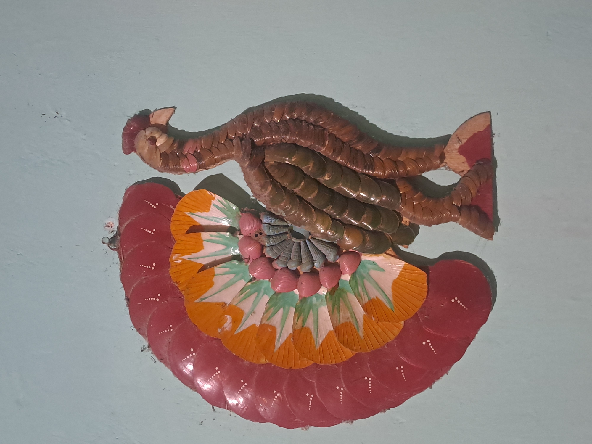 A decorative wall hanging of a peacock is affixed to a light blue wall in Sumberejo, Indonesia. The peacock is constructed from various materials, including shells and possibly dried leaves, and is depicted in profile facing right. Its tail fan is fanned out, displaying concentric layers of red, orange, and green, with the red outer layer adorned with small white dots. The body and neck of the peacock are formed from numerous small, brown, overlapping pieces, giving it a textured appearance. The head is detailed with a comb-like crest and a beak. The bird stands on a base made of wood painted red and brown. The background wall shows some minor imperfections and a visible hook at the top left, from which the artwork is hung.