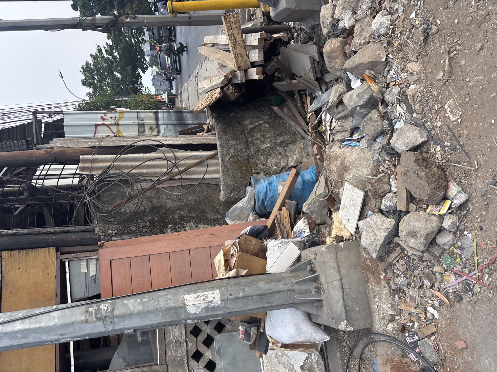 The image depicts a cluttered sidewalk in Bekasi, Indonesia, characterized by significant debris and discarded materials. A large, gray metal pole, possibly a utility pole, dominates the left side of the frame, anchored to a concrete base. Adjacent to it is a stack of wooden planks and other construction debris. Further into the scene, a blue tarp is visible among the rubble, along with various rocks, broken concrete, and scattered trash, including paper, plastic, and what appear to be discarded books or magazines. In the background, a street with moderate traffic is visible, with vehicles and a motorcycle on the road. Trees line the street, and a pedestrian walkway runs alongside it. The overall impression is one of disarray and neglect on the urban thoroughfare.