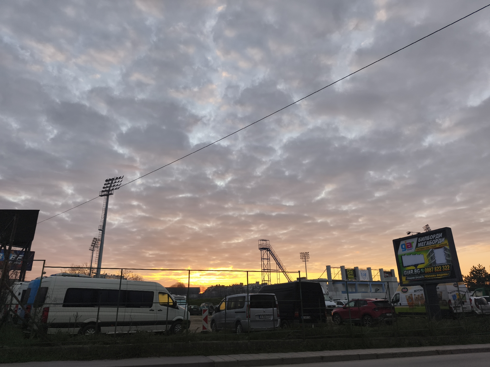 The image depicts a view in what appears to be an industrial or commercial area in Varna, Bulgaria, with a dramatic sky at sunset or sunrise. The lighting is orange and soft, casting an interesting glow on the clouds. The scene is somewhat cluttered with vehicles parked along a fence, including a white van, a silver van, a black van, and a red car. In the background is a stadium with floodlights, a metal staircase, and advertising billboards. One prominent billboard has Bulgarian text and contact information, "GIAR.BG 0887 822 322." The overall impression is one of urban infrastructure against the backdrop of a striking sky. A single wire stretches across the sky.