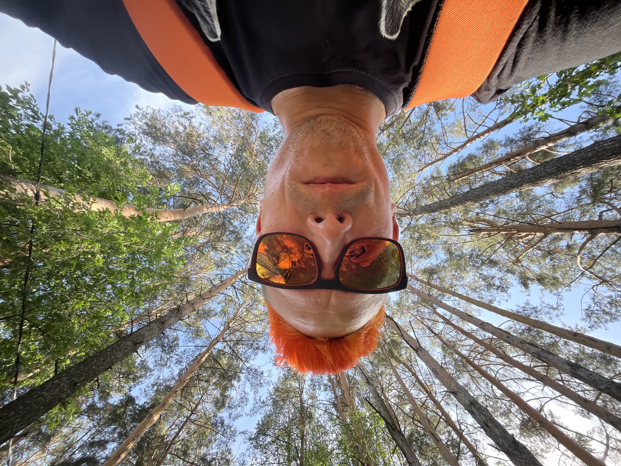 A low-angle view looking up at a person with an orange hairstyle, wearing black framed sunglasses with orange reflective lenses, and an orange harness over a black t-shirt. The background is filled with tall trees reaching up towards a blue sky with some clouds. The photo appears to have been taken in a forest or wooded area in Chaikovsky, Russia, based on the forest type. The person is upside down, possibly secured for a tree-climbing or aerial activity.