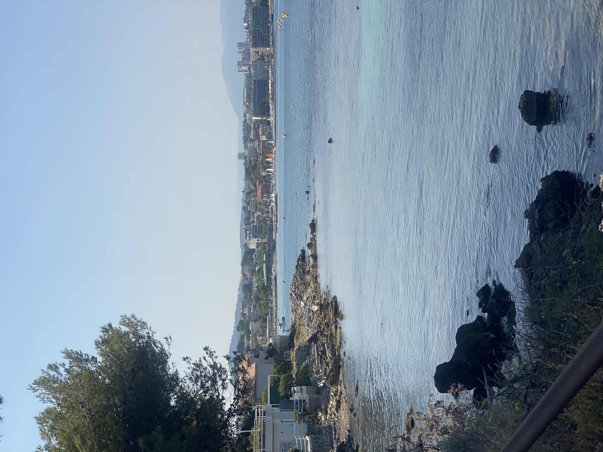 The image depicts a coastal view in Antibes, France. The foreground features a rocky shoreline with some vegetation and a dark railing. The water is a clear blue, with some darker rocks visible beneath the surface. In the midground, a white building with a terracotta roof is situated on the rocky coast, with a railing leading to it. A small white boat is visible on the water in front of the building. Further in the distance, a city skyline with several buildings, including apartment complexes and hotels, stretches along the coast. Sailboats with orange and red sails are dotted across the bay. Yellow buoys are scattered throughout the water. The sky is clear and blue with a slight haze, and distant mountains are faintly visible on the horizon. The scene is bathed in bright daylight, suggesting a sunny day.