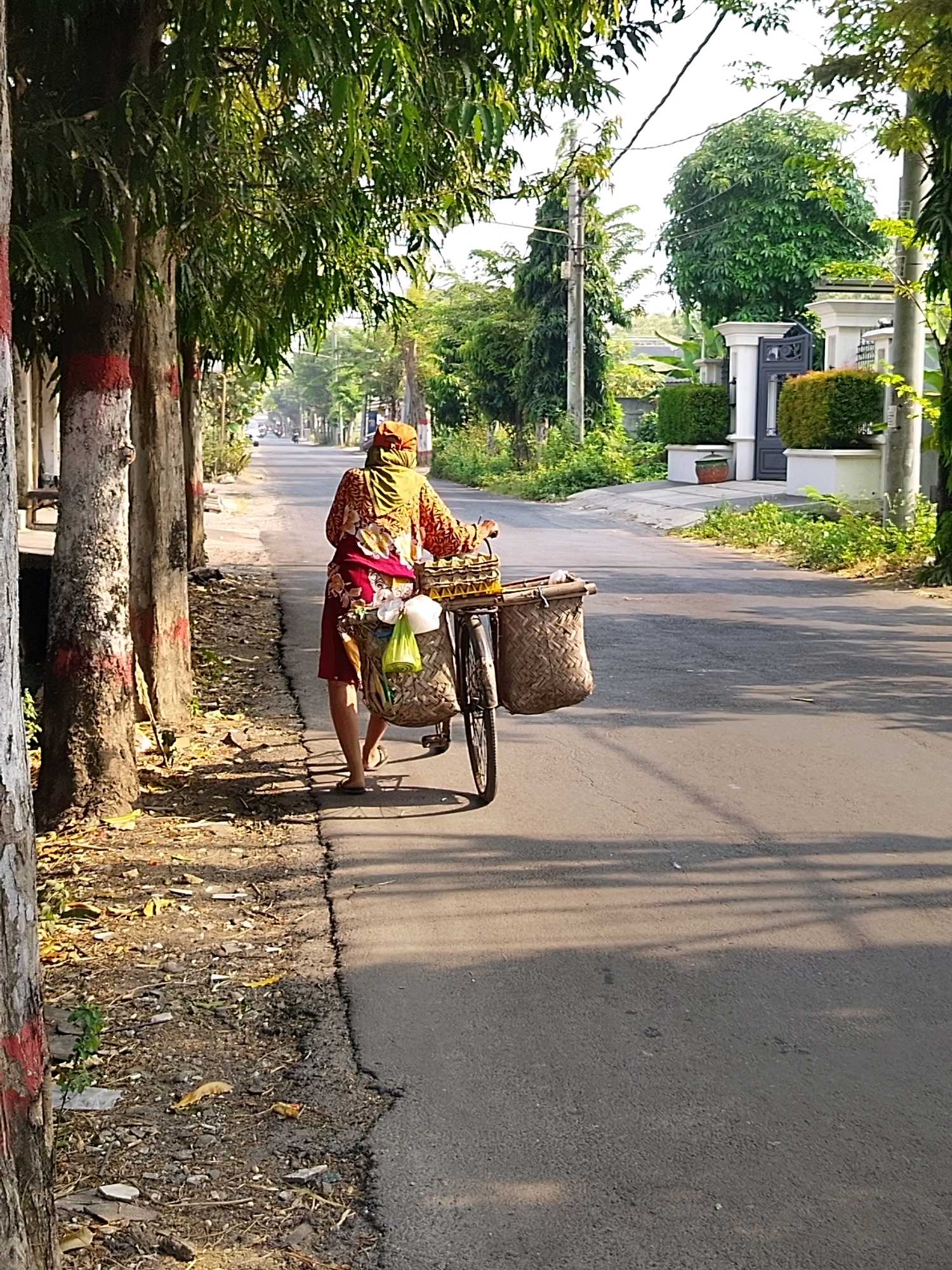 A person wearing a colorful patterned shirt, a red skirt, and a headscarf is pushing a bicycle on a paved road in Mojokerto, Indonesia. The bicycle has two baskets attached, one in front and one behind the rider. The front basket appears to contain yellow crates, and the rear basket has woven texture and a green plastic bag hanging from it. The person is walking on the left side of the road, next to a dirt path lined with trees. The trees on the left have red and white markings on their trunks. In the background, the road continues, with houses and greenery visible on the right side. The sun is casting long shadows across the road.