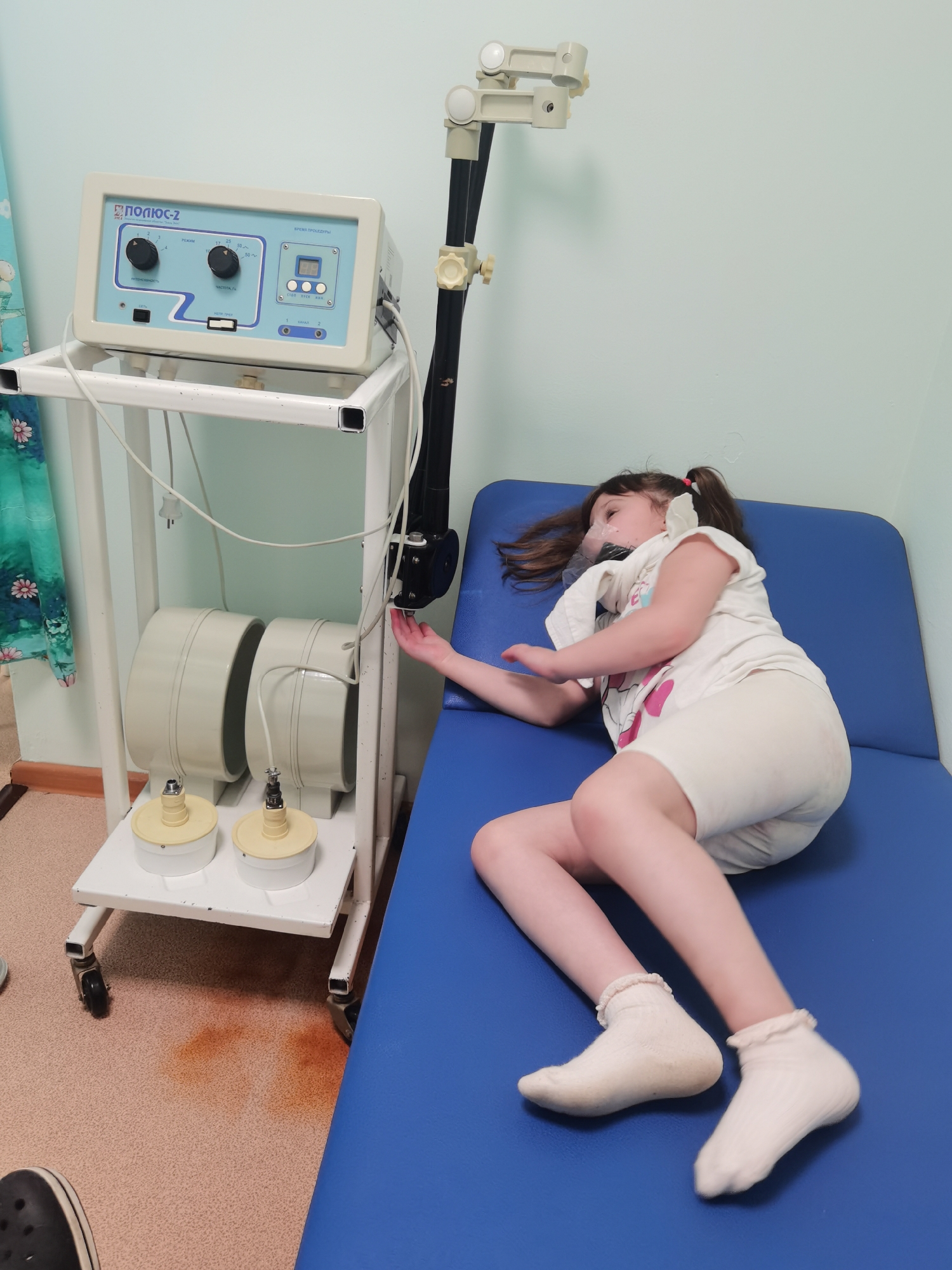 A young girl is lying on a blue examination table in what appears to be a medical facility. She has brown hair pulled back in pigtails with white scrunchies and is wearing a white t-shirt and white shorts. She is also wearing a medical face mask.
Next to the table is a medical device on a white rolling cart. The device has a control panel labeled "ПОЛЮС-2" in Cyrillic, and two large circular magnetic field emitters are on the lower shelf of the cart. A movable arm extends from the cart, positioning a dark sensor near the girl. There are brown stains on the floor near the cart. A teal curtain is visible in the background, and the walls appear to be a light blue or green color. A black Croc style shoe can be seen in the lower left corner. The location is Perm, Russia.