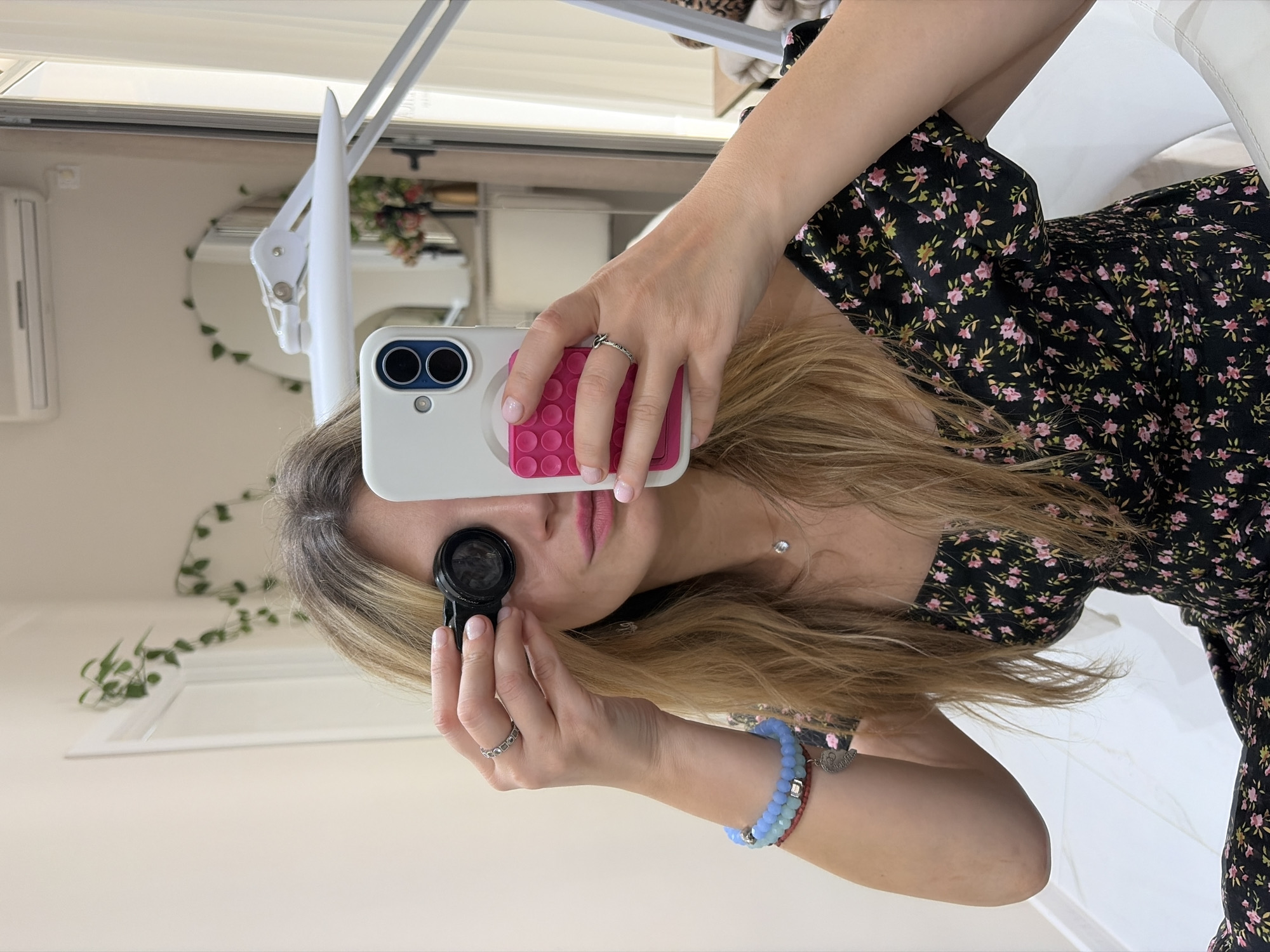 A woman is shown from a close-up, slightly tilted perspective. She is wearing a black dress with small pink floral patterns and has long, blonde hair. She is holding a smartphone in her right hand, which has a white case and a pink, textured grip on the back. She is using the phone to take a close-up photo of her eye with a small, round device attached to the phone's camera. The woman is wearing a bracelet with blue and silver beads on her left wrist. The background shows a brightly lit room with a mirror, a light fixture, and an air conditioning unit. The location in the city of Budva, Montenegro, is not discernible from the visual data provided.