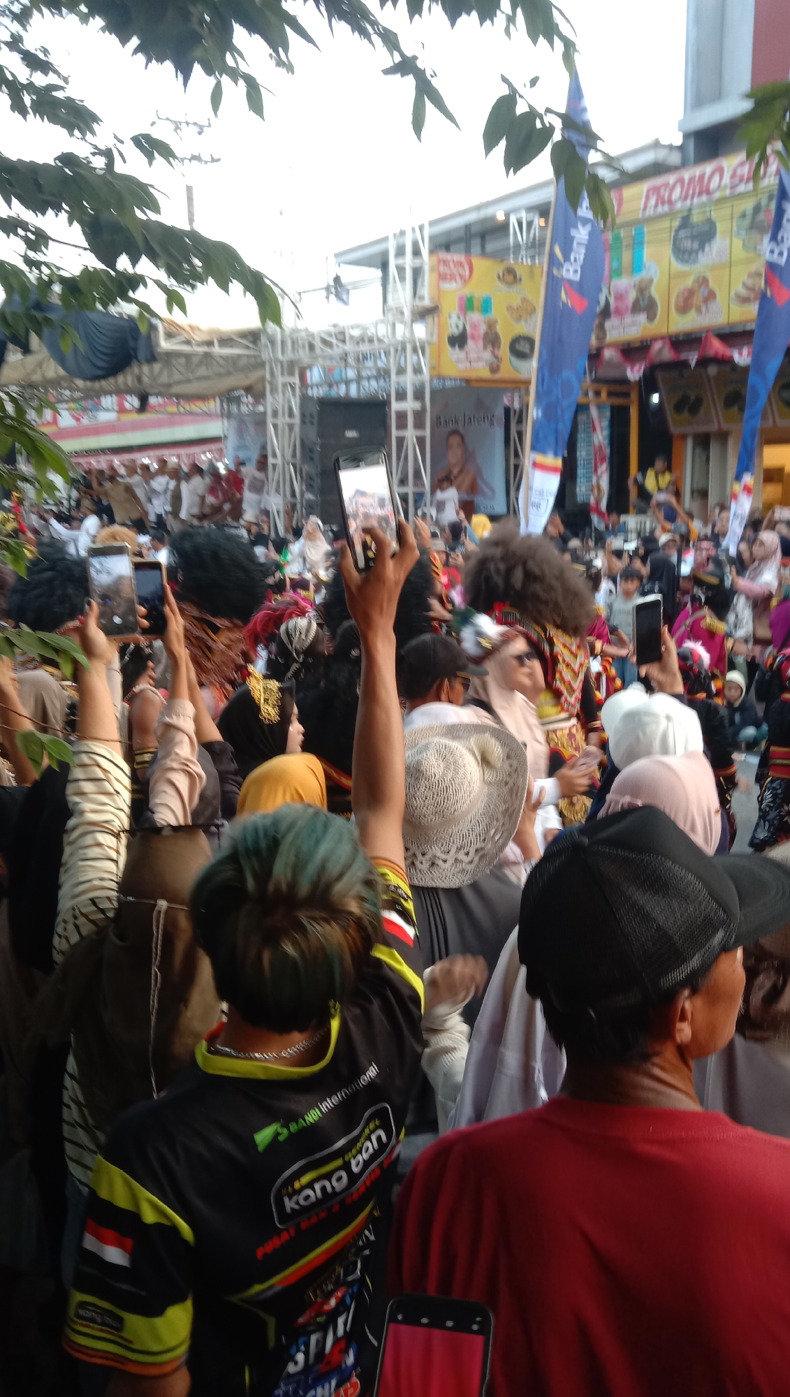 The image shows a crowd of people gathered outdoors, likely for a cultural event or festival. In the foreground, several individuals are holding up smartphones to capture the scene, suggesting a performance or procession is taking place. One person on the left is wearing a black and yellow shirt with text that includes "kang ban" and "International." To their right, a person in a wide-brimmed straw hat is visible. In the background, there's a stage or performance area with a large screen or banner displaying advertisements, including "Bank Jateng" and "PROMO SHOP." Several individuals in elaborate costumes, possibly dancers or performers, are visible within the crowd and on or near the stage. The atmosphere appears lively and crowded. The location is identified as Jatisawit, Indonesia.