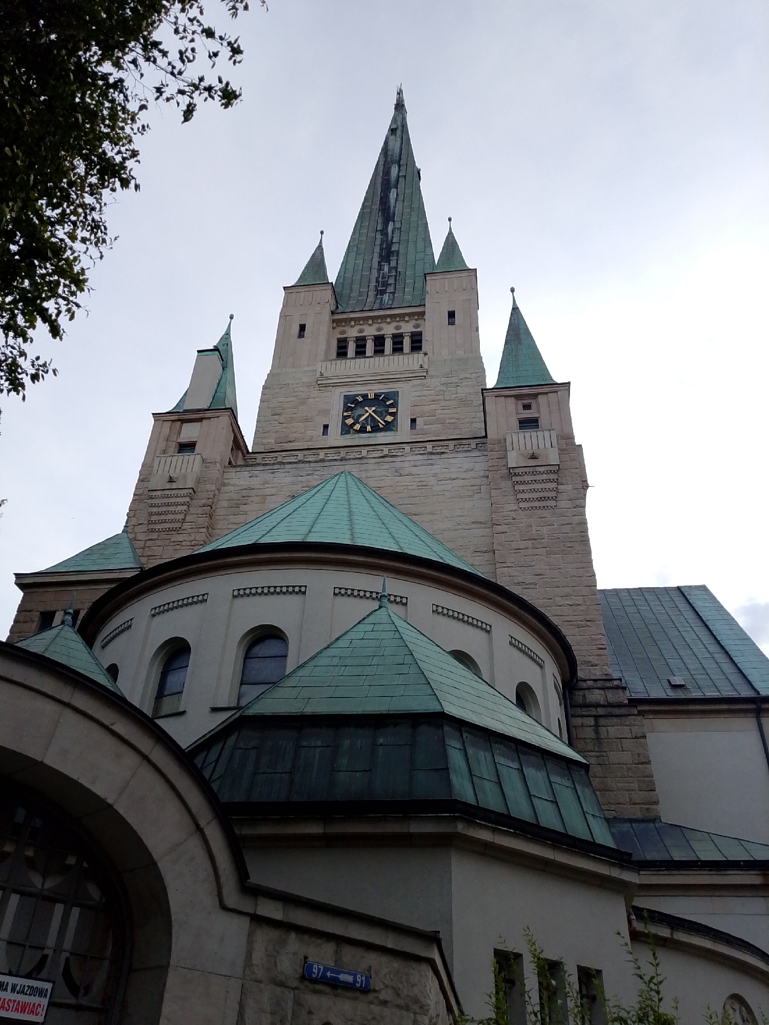 The image shows a low-angle view of the Holy Trinity Church in Wrocław, Poland. The church features a tall, square tower topped with a pointed spire, flanked by smaller spires. The tower has a clock face with Roman numerals and golden hands. The building's exterior is constructed from light-colored stone and concrete, with several arched windows and green, tiled roofs. In the foreground, a curved stone archway frames a portion of the building and a sign with Polish text is visible. There are trees and foliage partially obscuring the lower left side of the church. The sky is overcast.