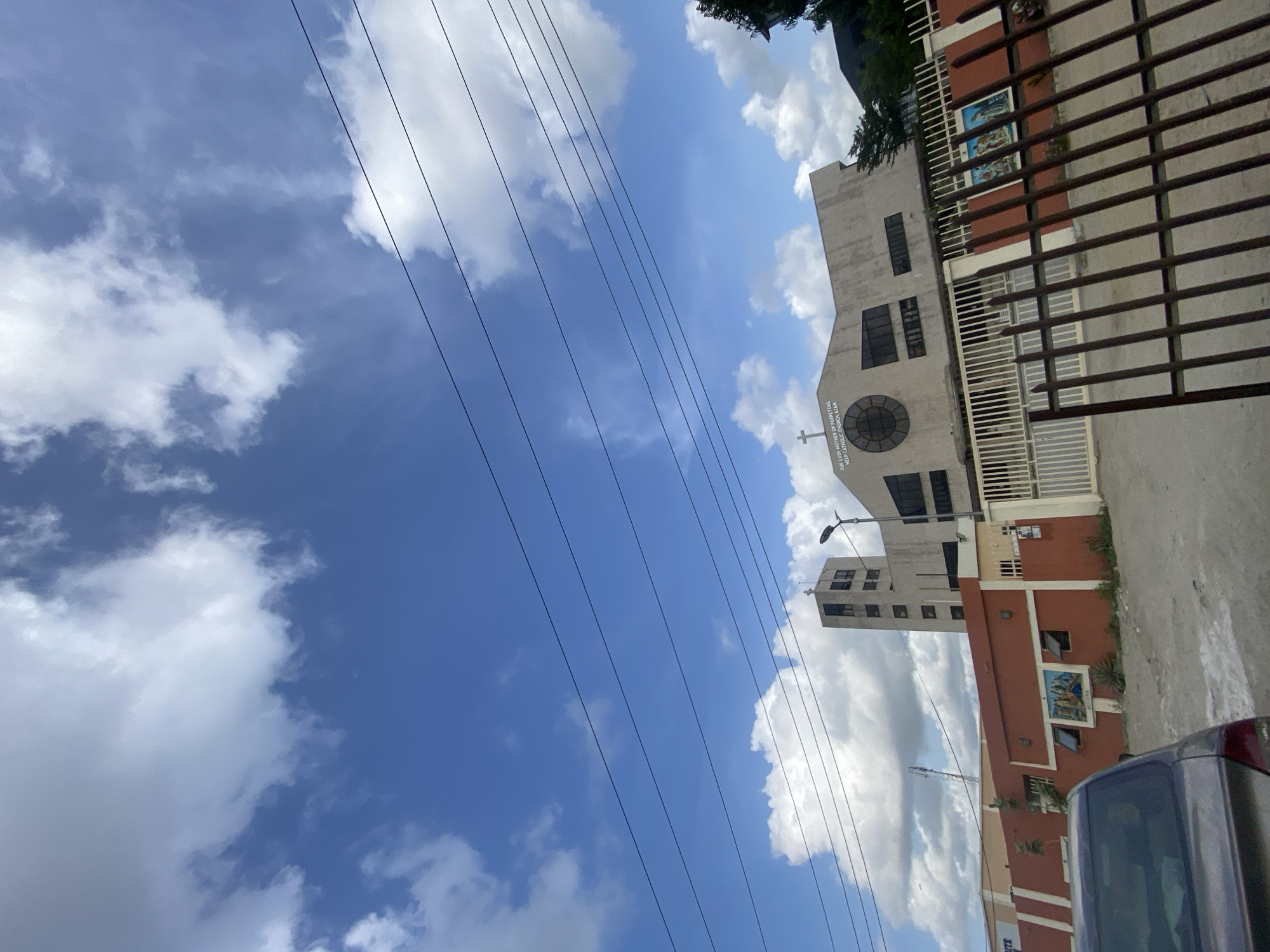The image shows a low-angle view of the Our Lady Mother of Perpetual Help Catholic Church in Lekki, Nigeria. The church building is visible, with a tower, and exterior elements such as windows and architectural details are partially obstructed by a dark gate. The sky is prominent, filled with clouds and intersected by power lines. Part of a vehicle, possibly a car, appears in the lower right corner. The image is rotated, presenting a tilted perspective of the scene.