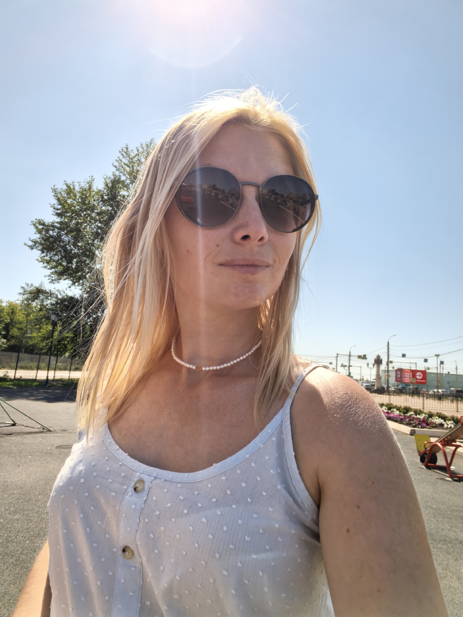 In Sterlitamak, Russia, a woman with blonde hair and sunglasses takes a selfie outdoors on a sunny day. She is wearing a light blue, polka-dotted tank top and a pearl necklace with a small, heart-shaped pendant. The background shows a clear blue sky, a tree, a lamppost, a fence, and some playground equipment. The woman is positioned in the foreground, with her face turned slightly to the right.