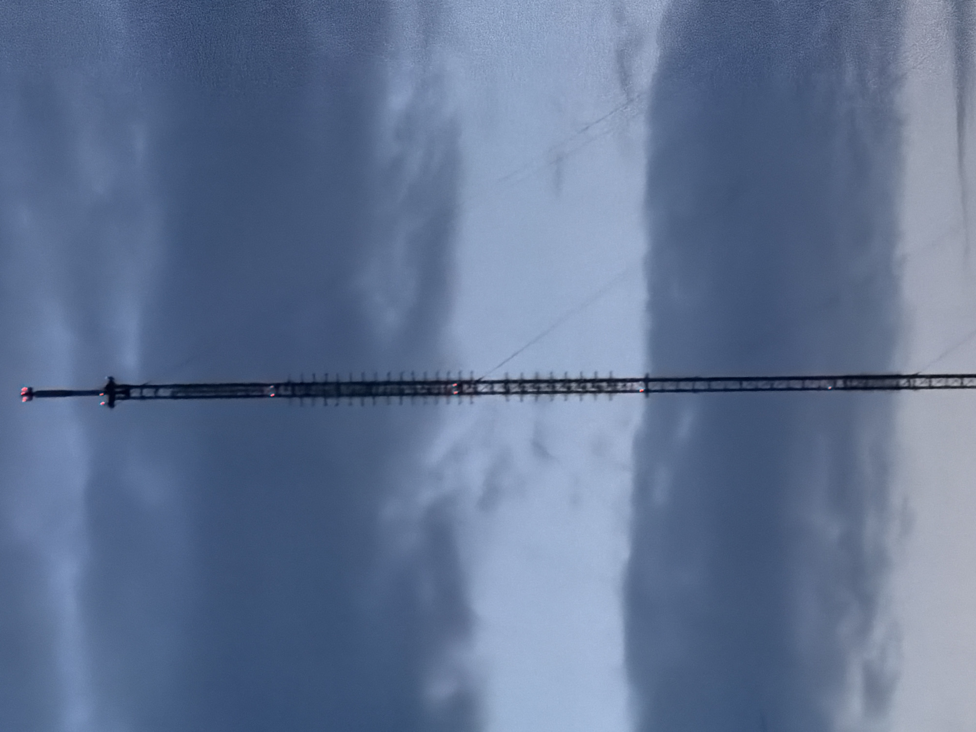 The image shows a tall transmission tower against a cloudy sky. The tower appears to be a lattice structure with multiple segments, and a red warning light is visible at its top. The clouds vary in color and density, suggesting an overcast day. There is no information to determine the exact location of this tower in Malinovka, Russia. No people, subjects, objects, actions, or interactions are visible besides the tower and sky.