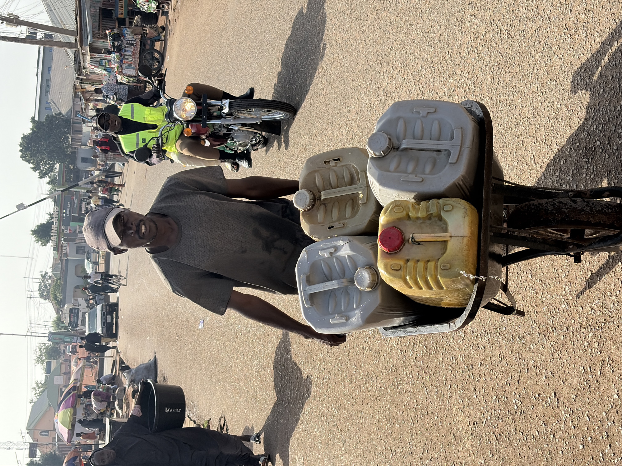 In Jos, Nigeria, a man in a grey t-shirt and cap stands on a paved street, facing the camera with a smile. He is pulling a wheelbarrow filled with four large, rectangular plastic jerrycans. One jerrycan is yellow with a red cap, while the others are light grey. A motorcyclist wearing a bright yellow vest is passing by in the background. The street shows signs of market activity with vendors and pedestrians. Various buildings and vehicles line the street, and sunlight casts strong shadows across the pavement. A woman is passing in a black abaya carrying a black bucket.