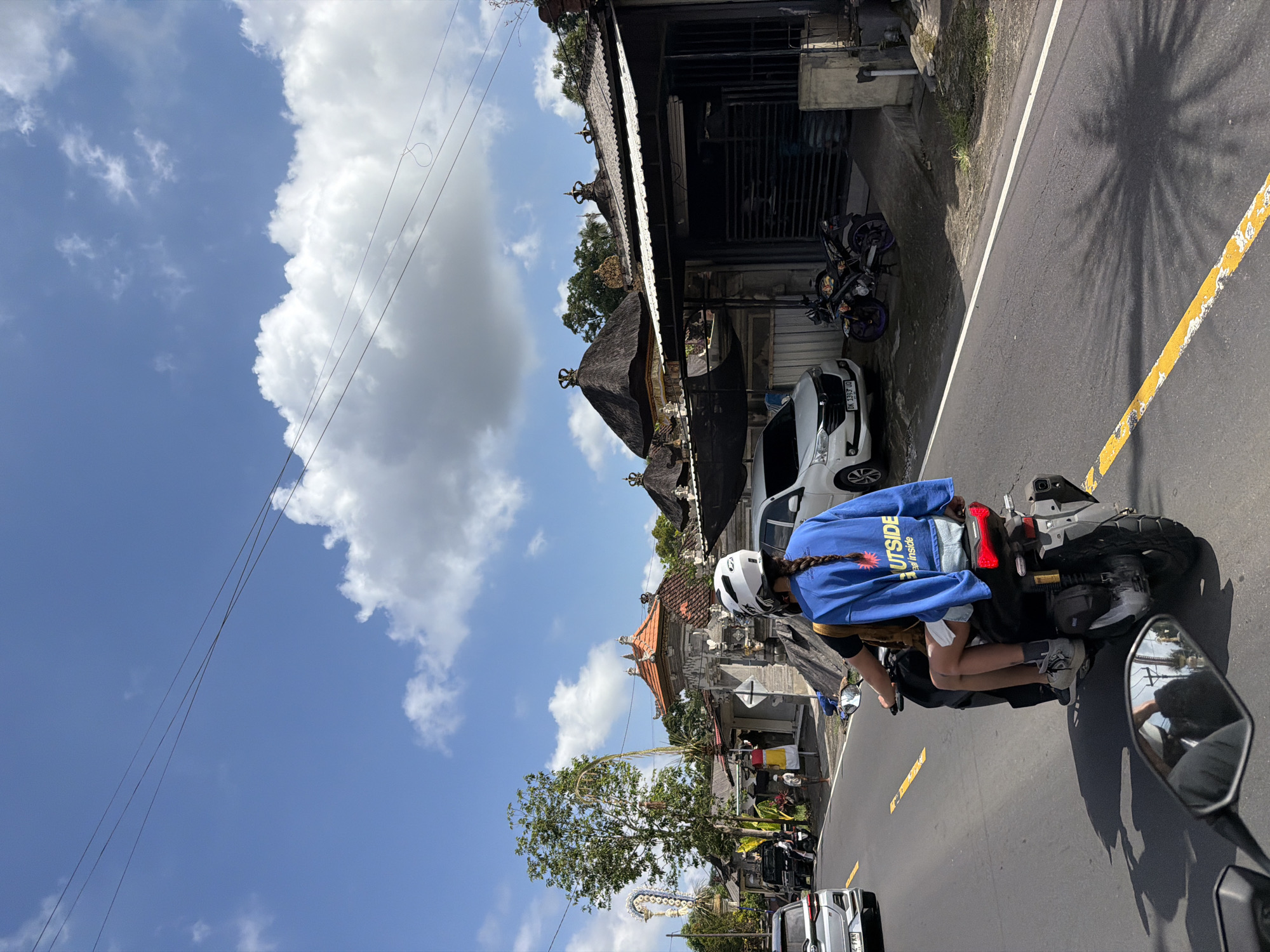 In Tampaksiring, Indonesia, two individuals ride a scooter along a paved road under a bright blue sky with scattered clouds. The person in the passenger seat, wearing a blue sweatshirt with the word "OUTSIDE" in white lettering and denim shorts, has their hair in a braid. They are also wearing a white helmet. The driver, partially obscured, wears a dark shirt and is also on the scooter. Several vehicles, including a white car and a motorcycle, are parked on the side of the road. Buildings with traditional Balinese architecture, characterized by their peaked roofs and ornate facades, line the street. The scene captures a moment of daily life in a tropical urban environment, with lush green trees visible in the background.