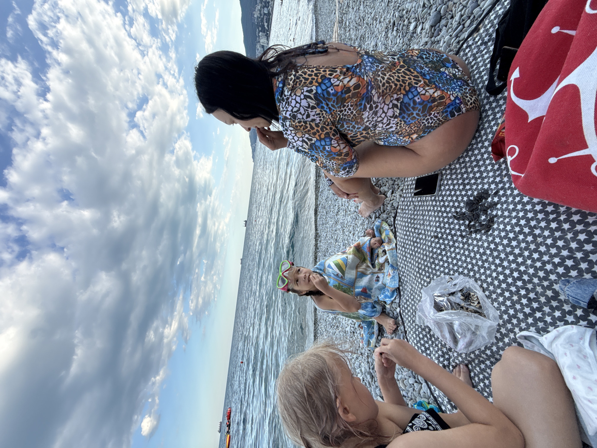 The image depicts three individuals at a beach, with the sea and a cloudy sky in the background. The scene appears to be set in Yalta, Ukraine.
In the foreground, a young girl with blonde hair is sitting and looking down, seemingly engaged with something in her hands. To her left and slightly behind her, another child, possibly a girl, is also sitting, wrapped in a blue and white towel. She is wearing green goggles and is looking up and to the left.
In the background, an adult with long dark hair is crouching and looking down. She is wearing a colorful, patterned dress with a leopard-like print. The individuals are sitting on a patterned blanket spread out on a pebble beach. A red towel with white anchor patterns is visible on the right side of the image. A clear plastic bag filled with what appears to be sunflower seeds is also present on the blanket.
The sky is filled with a mix of blue sky and dense clouds, suggesting it might be a partly cloudy day. The sea is calm, with gentle waves. The overall impression is a relaxed, casual outing at the beach. No visible text is present in the image.