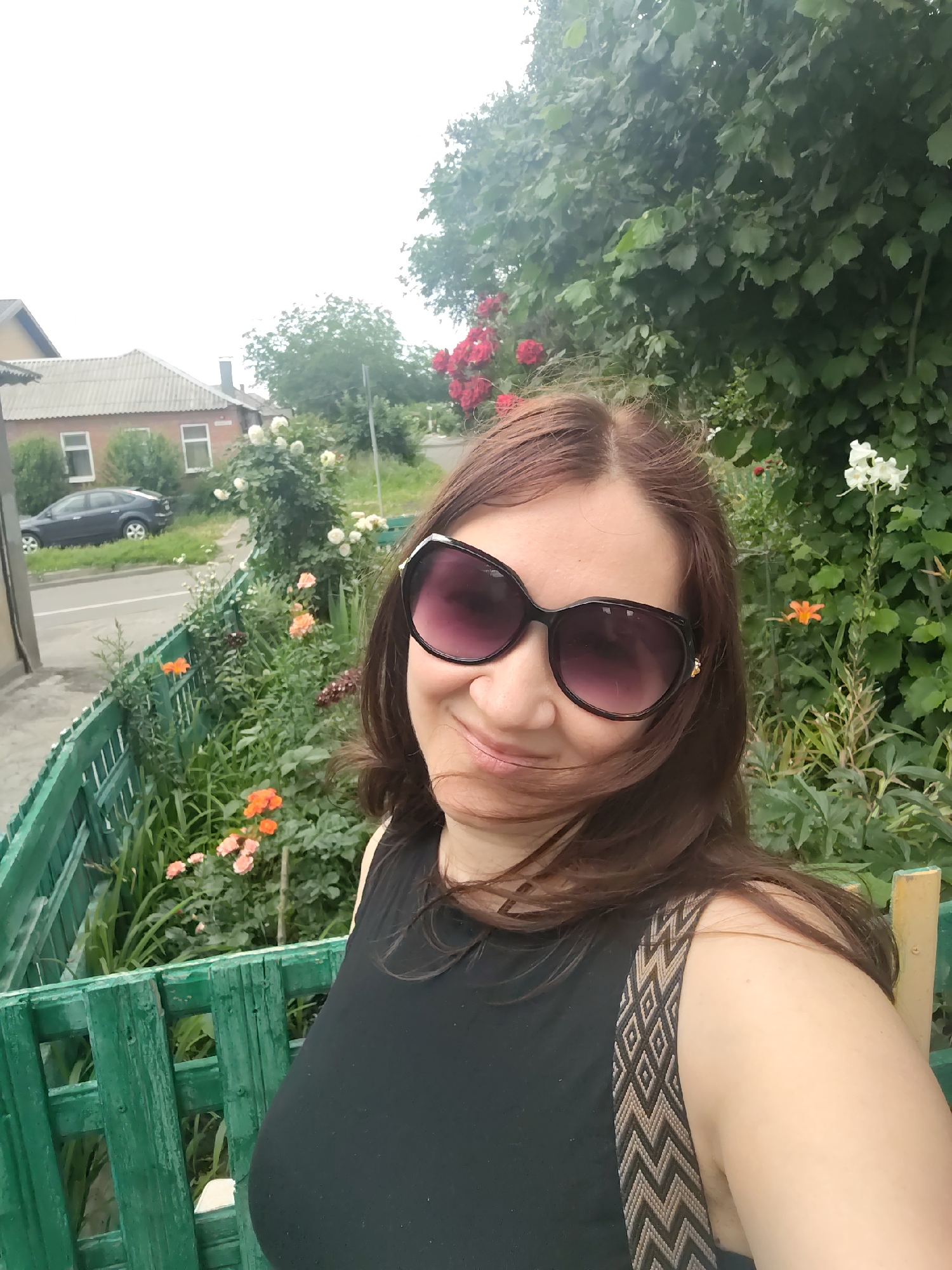 This is a selfie of a woman with long, brown hair, wearing large, dark-rimmed sunglasses and a black tank top with a patterned strap. She is smiling and looking at the camera. The background shows a garden with various flowers, including red roses, white roses, and orange flowers. A green wooden fence runs along the front of the garden. Behind the garden, there are houses with gray roofs and windows, a road with a dark-colored car, and lush green trees and foliage. The sky appears overcast, suggesting daytime. The overall scene depicts a casual moment outdoors, likely in a residential area, with a focus on the woman and the blooming garden. The image appears to be taken in Rostov-on-Don, Russia. There is no visible text.