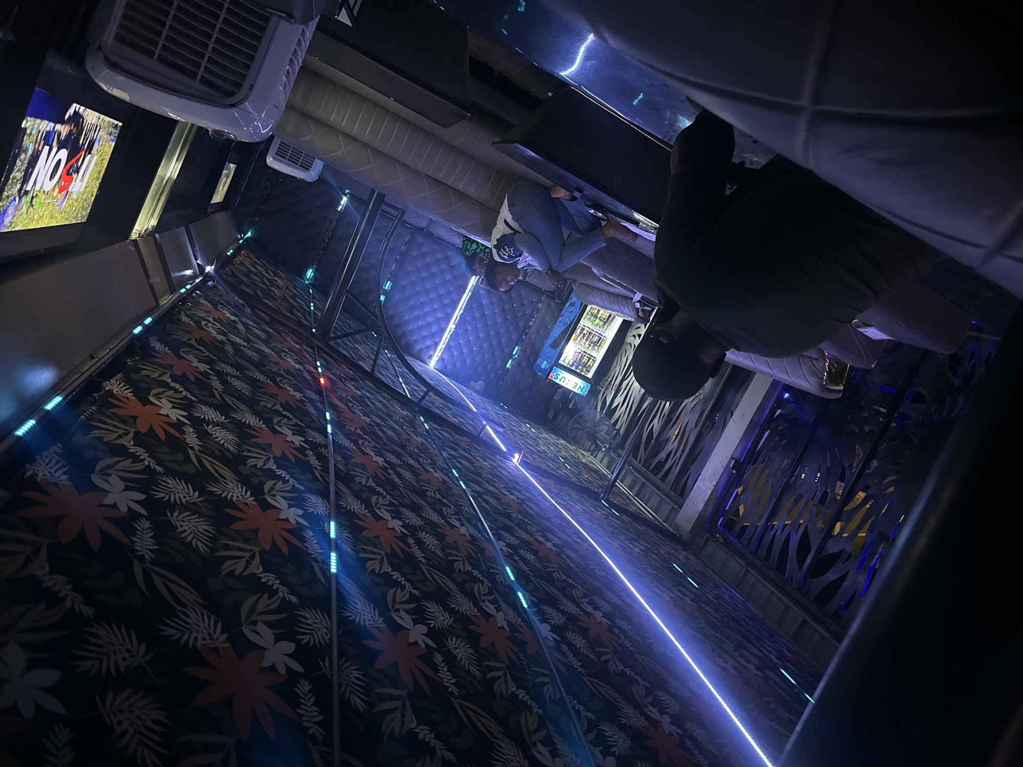 The image depicts the interior of a karaoke room, possibly in Tumfure, Nigeria. The room features patterned carpeting with floral motifs and strip lighting along the walls. A lounge area with cushioned seating and a table is visible. Several individuals are present, one of whom is wearing a dark top and appears to be seated, while another is wearing blue jeans and a white top. A display with the word "NO" is visible in the background, along with a beverage display labeled "NEXUS". An air conditioning unit is mounted on the ceiling. The room has decorative metal panels and a generally dim, blue-toned lighting scheme.