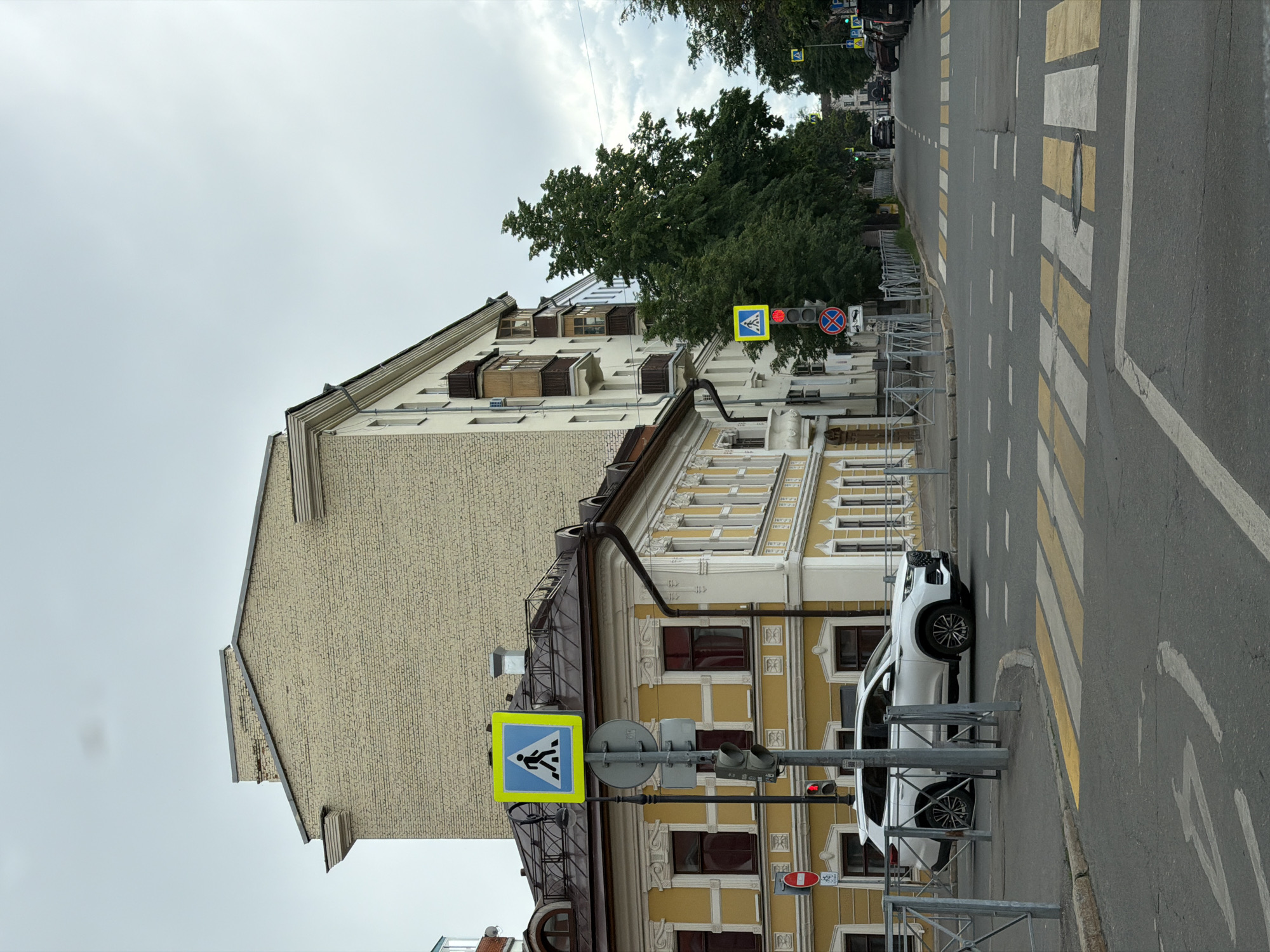 The image shows a street scene in Kazan, Russia. The primary focus is on a street crossing, indicated by a blue and yellow pedestrian crossing sign. A traffic light is visible, showing a red light. A white car is parked near the pedestrian crossing. The architecture of the building near the crossing includes a yellow facade and windows with ornate frames. The street features a crosswalk marked with alternating yellow and white stripes. Further down the street, there are other vehicles and trees, suggesting an urban environment. The sky is overcast.