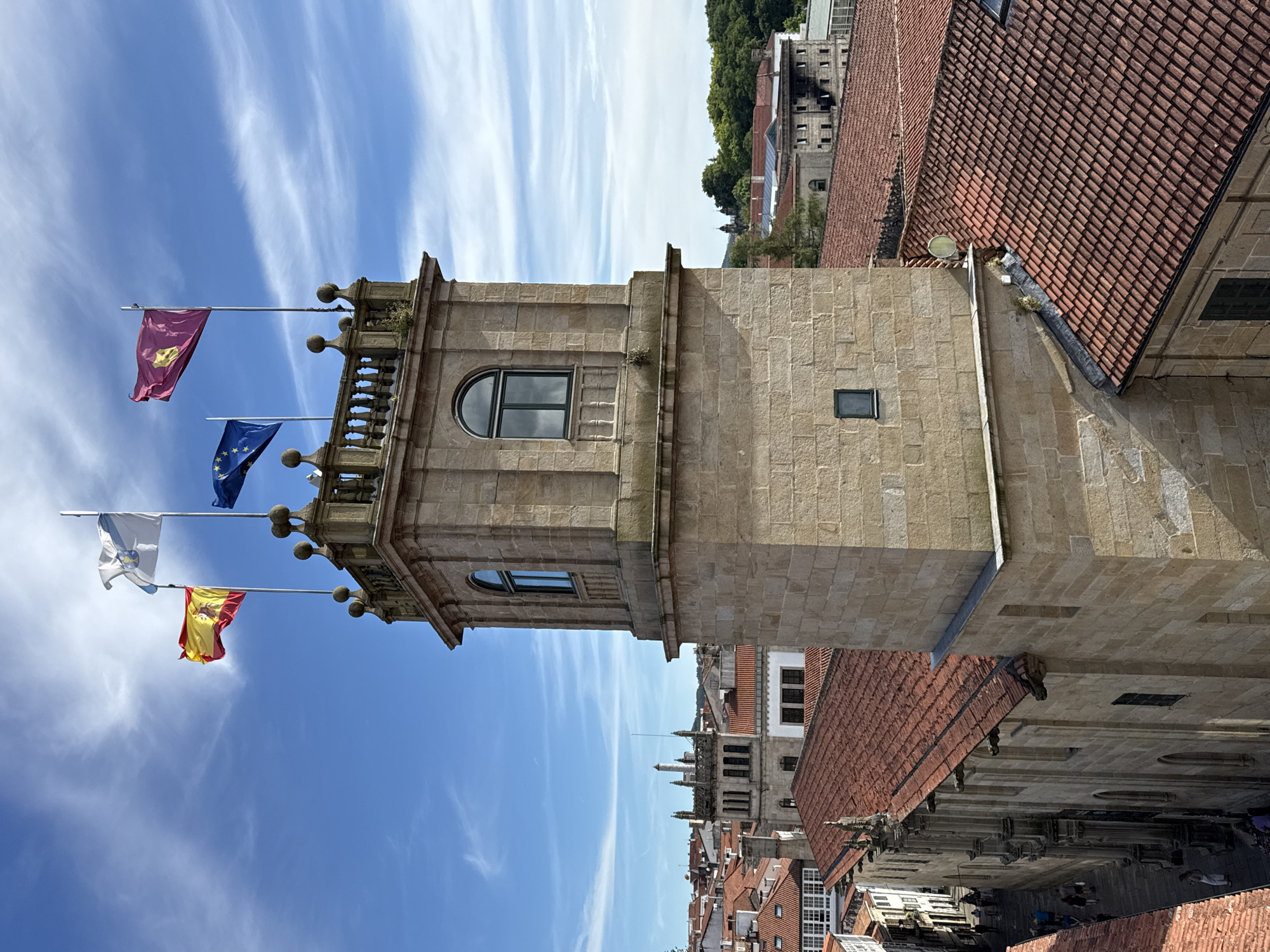 The image shows a view from above in Santiago De Compostela, Spain, focusing on a stone tower adorned with several flags. The flags visible are the Spanish flag, the European Union flag, a white flag with a star and possibly a symbol, and a maroon flag with a yellow emblem. The tower is constructed from large, light-colored stones and features arched windows with dark panes and decorative stonework. Below the tower, red-tiled rooftops dominate the scene, indicative of traditional Spanish architecture. On the left side of the image, a narrow street is visible with several people walking along it. The sky is blue with scattered white clouds. The overall scene captures a historic urban environment.