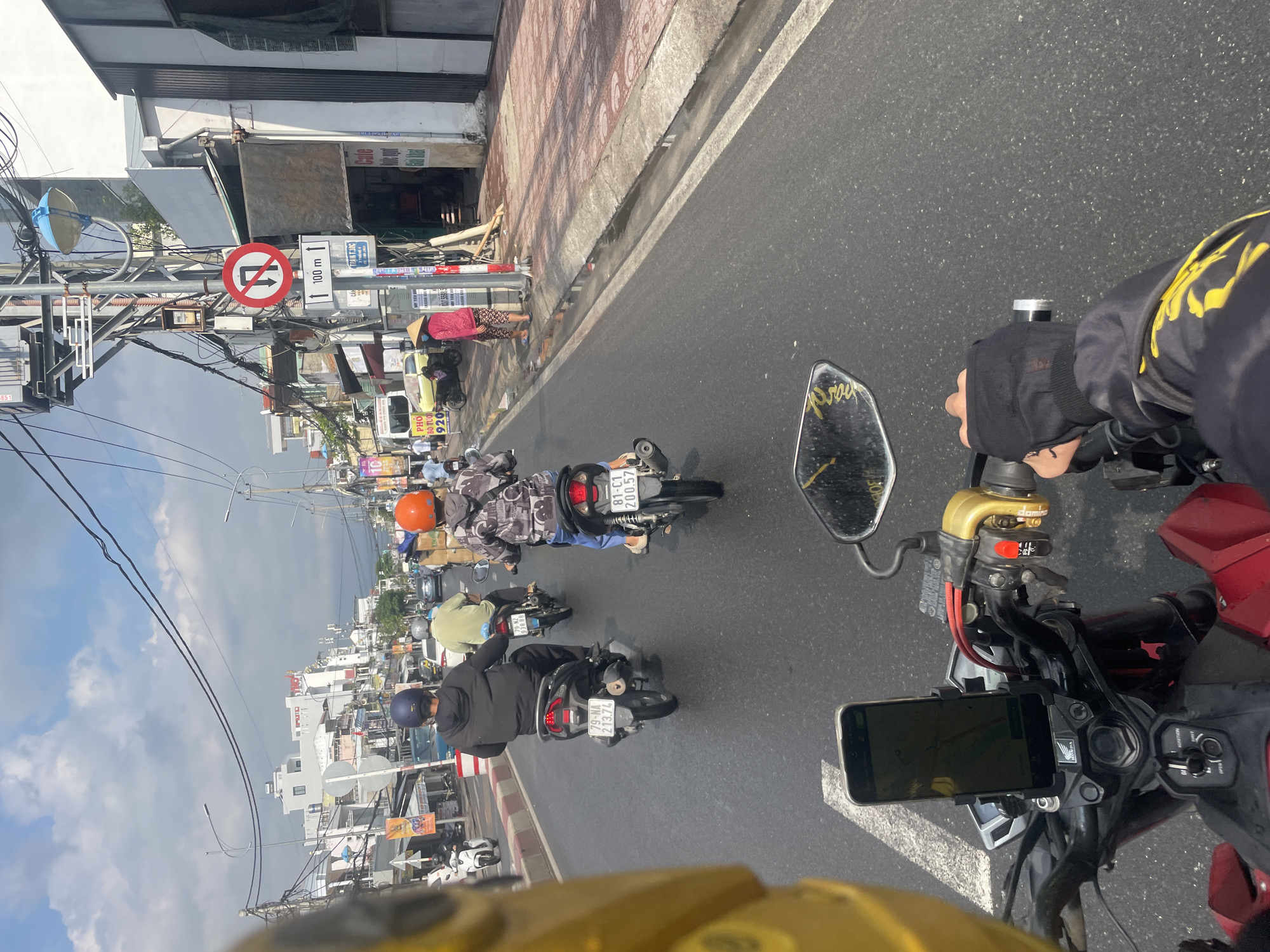 The image shows a first-person perspective from a motorbike rider in what appears to be a street in Bac Nha Trang Ward, Vietnam. There are several other motorbikes ahead, moving in the same direction, each with a single rider. In the foreground, the handlebars, side mirror, and a phone mount of the rider's motorbike are visible. A sign in the distance with a traffic restriction symbol and "100 m" suggests a prohibition of a certain type of turning within 100 meters. Buildings are on the sides of the road, and the sky is partly cloudy. The general impression is of a busy street with active motorbike traffic in an urban setting.