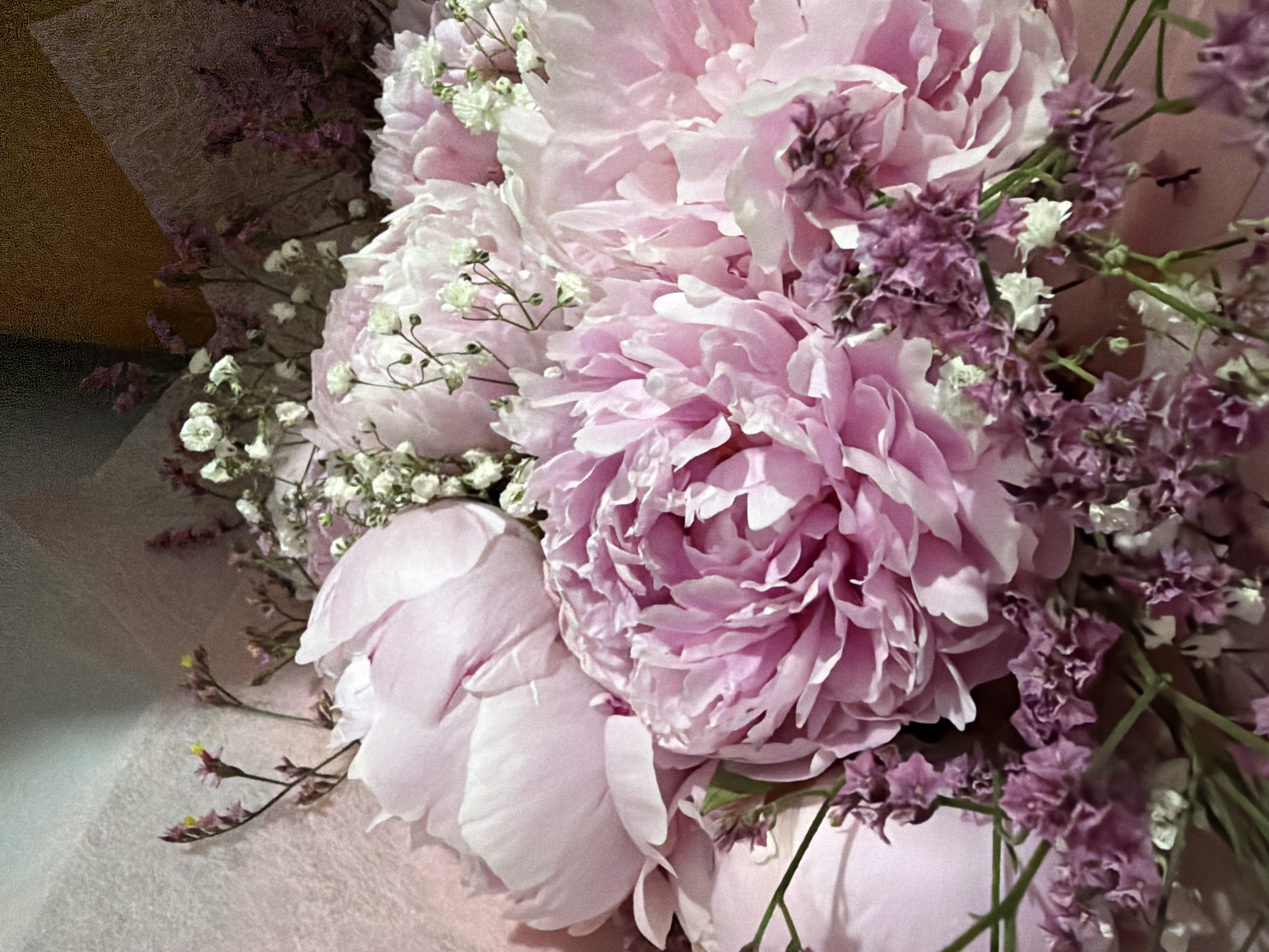 A close-up shot of a bouquet of pink peonies and delicate white baby's breath flowers is presented. The peonies, in various stages of bloom, display soft, ruffled petals in shades of light pink and white. The baby's breath flowers, small and clustered, add a whimsical touch to the arrangement. Some dried purple flowers with thin stems are also interspersed throughout the bouquet, adding texture and a contrast in color. The background is softly blurred, with hints of textured pink wrapping paper and a darker, warmer tone that could be wood or fabric. The lighting is soft and diffused, highlighting the delicate beauty of the flowers. The scene is static and appears to be a still-life arrangement.