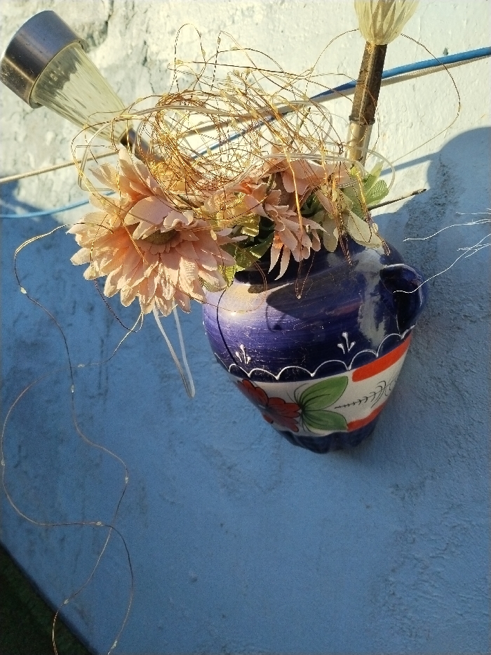 A blue and white wall-mounted vase holds peach-colored flowers and decorative copper-colored wire. A solar-powered garden light, with a silver top and clear conical lens, is positioned to the left. A second, taller, solar light with a similar design is visible behind the floral arrangement. The background is a light blue textured wall, casting shadows and highlighting the vase.