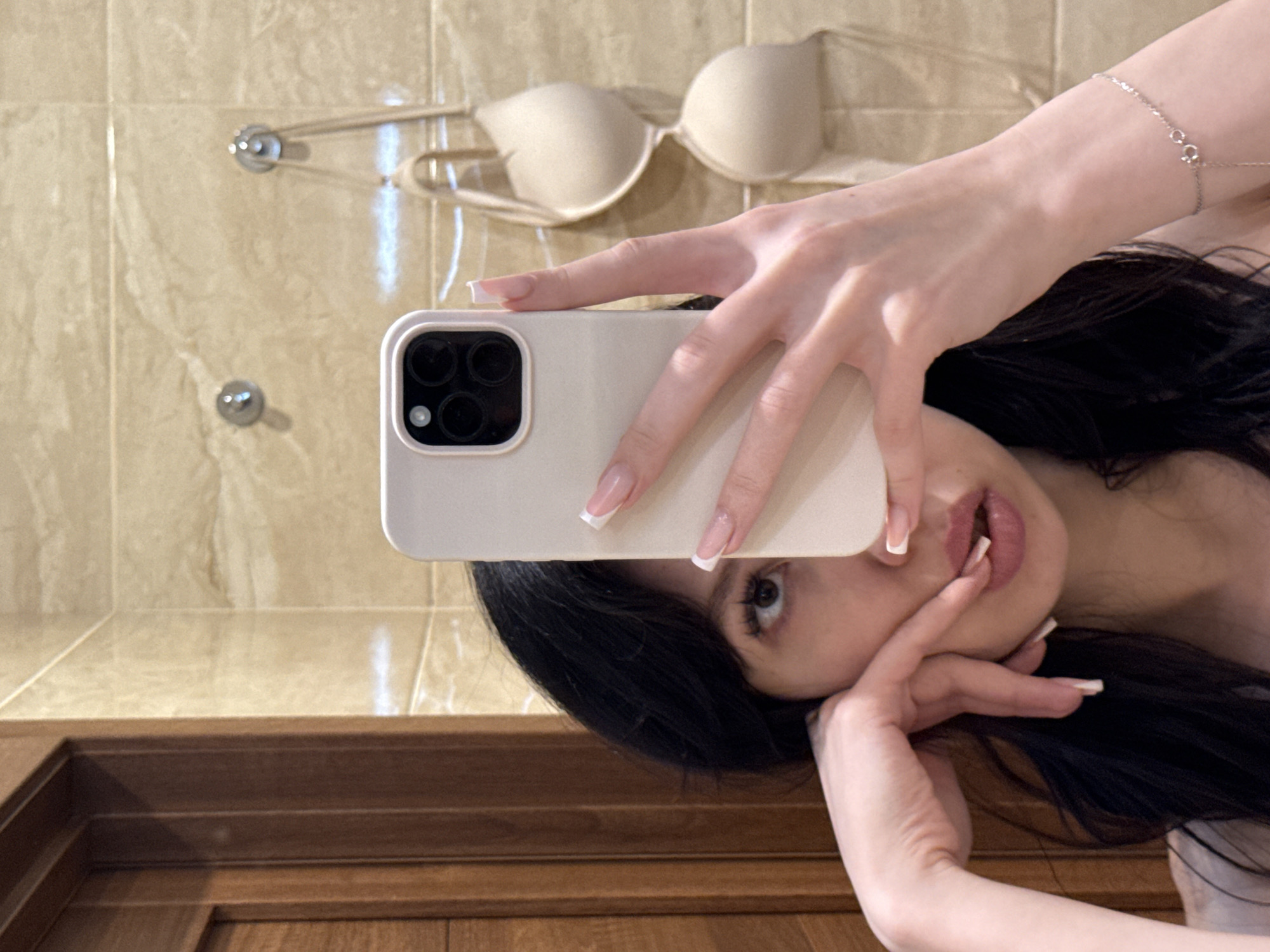 A young woman is taking a selfie in a bathroom. She has dark hair and is wearing a light-colored top. Her left hand is on her cheek and her right hand is holding up a phone to take the picture. A beige bra is hanging on the wall behind her. The bathroom has tiled walls. The image is likely taken in Khimki, Russia.