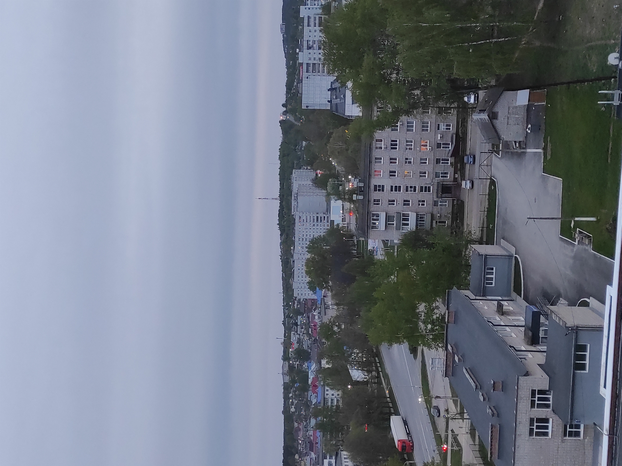 The image is a vertically oriented, high-angle, eye-level shot of a cityscape in Chaikovsky, Russia. The sky is overcast, casting a soft, diffuse light across the scene.
Several multi-story apartment buildings are visible, mainly constructed in a blocky, Soviet-era style. These buildings are primarily gray, with rows of windows. Some windows appear illuminated, suggesting it is dusk or early evening. A few trees with green foliage are scattered throughout the scene, partially obscuring some of the buildings. A road runs through the middle of the image, and a long-haul truck is seen stopped at a red traffic light. A large commercial building is positioned in the lower right corner of the image.