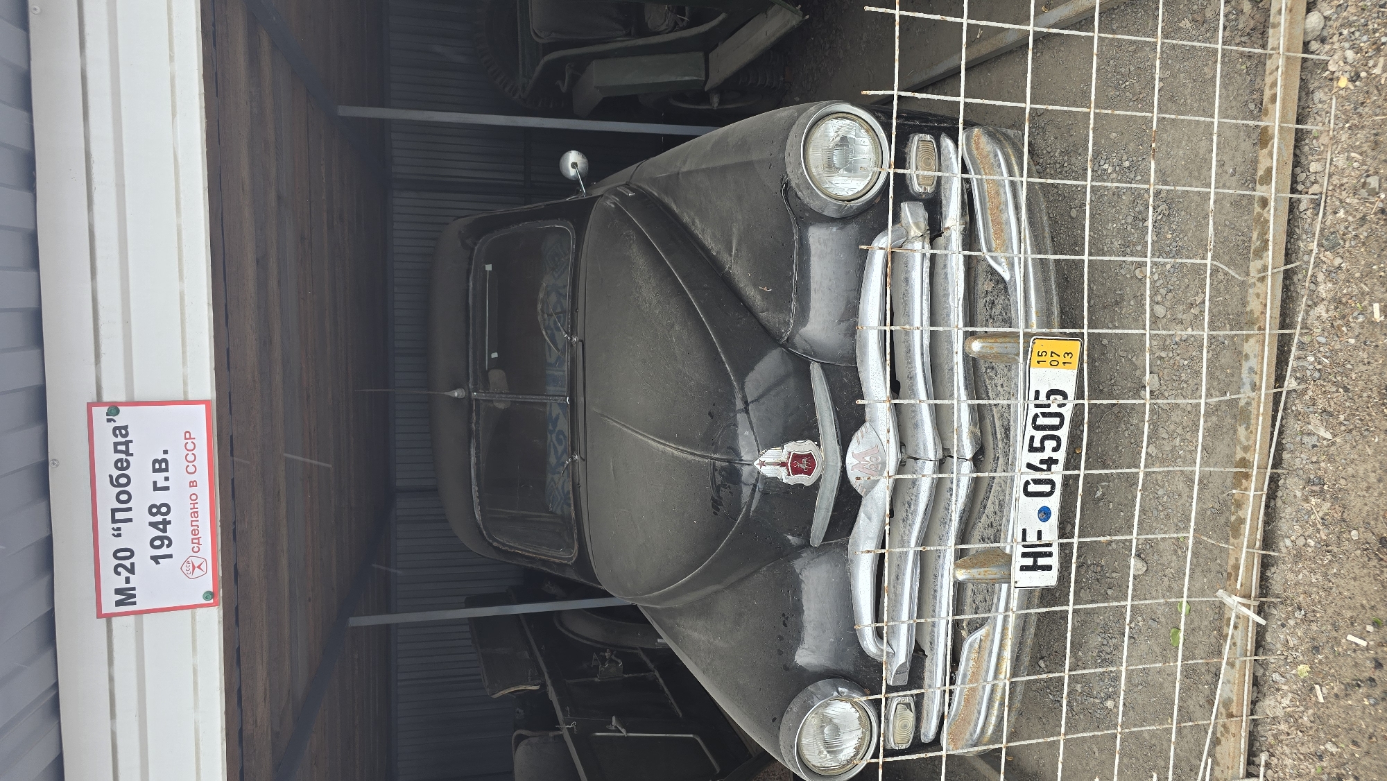 The image shows a dusty, black Soviet-era car, identified as an M-20 "Pobeda" from 1948, parked under a metal roofed shelter. The car has a chrome grille, round headlights, and a red emblem on its hood. A license plate reading "HF 04505" is visible on the front. A small yellow sticker with "15 07 13" is affixed to the license plate. The car is behind a wire fence. Other vehicles, appearing to be vintage military jeeps or trucks, are partially visible in the background to the left and right of the M-20. The ground is covered in gravel and dirt. A sign above the car reads "M-20 'Победа' 1948 г.в. сделано в СССР" which translates to "M-20 'Victory' 1948 year of manufacture. Made in the USSR". The location is stated to be in the city of Bol'shoi Mashlyak, Russia.