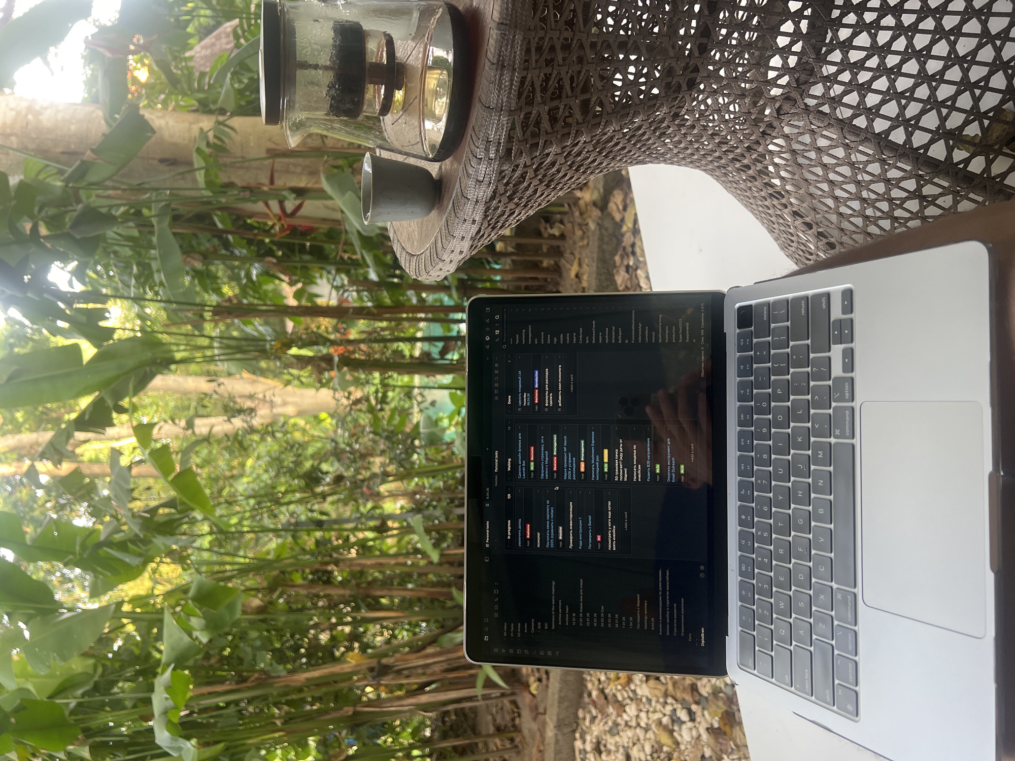 The image shows a laptop on a table in an outdoor setting, possibly a garden or patio, in Kerobokan, Indonesia. The laptop screen displays what appears to be a project management software interface in Russian. A coffee brewing apparatus and a small cup sit on the same table. The environment includes lush green foliage and plants. No people are visible.