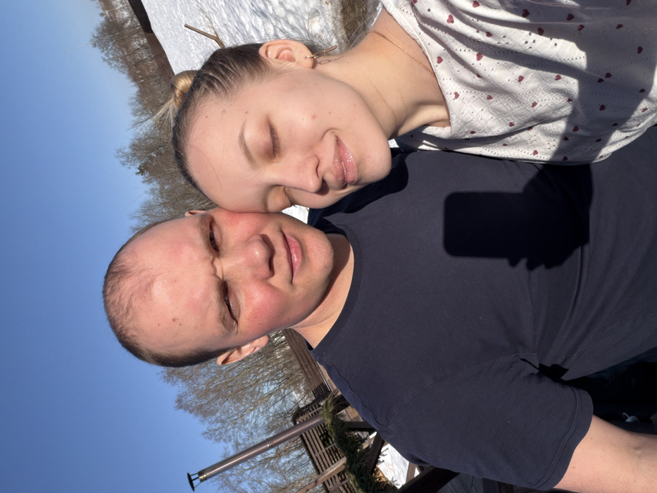 This image captures two individuals, a male and a female, posing outdoors on a clear, sunny day in Shipulino, Russia. The male, positioned on the left, has fair skin with a ruddy complexion, light brown hair with a receding hairline, and wears a dark blue short-sleeved t-shirt. He looks directly at the camera with a furrowed brow and slight squint. The female, on the right, leans her head against the male, her eyes closed, and a subtle smile on her face. She has light brown hair tied back, wears a white long-sleeved top adorned with small red heart patterns, small hoop earrings, and a thin gold chain. The background features extensive patches of snow-covered ground, several bare deciduous trees, and a clear blue sky. A tall, dark-colored ventilation or chimney pipe with a cap, alongside a wooden fence structure, is visible to the left of the male. The distinct shadow of a smartphone is cast upon the male's t-shirt.